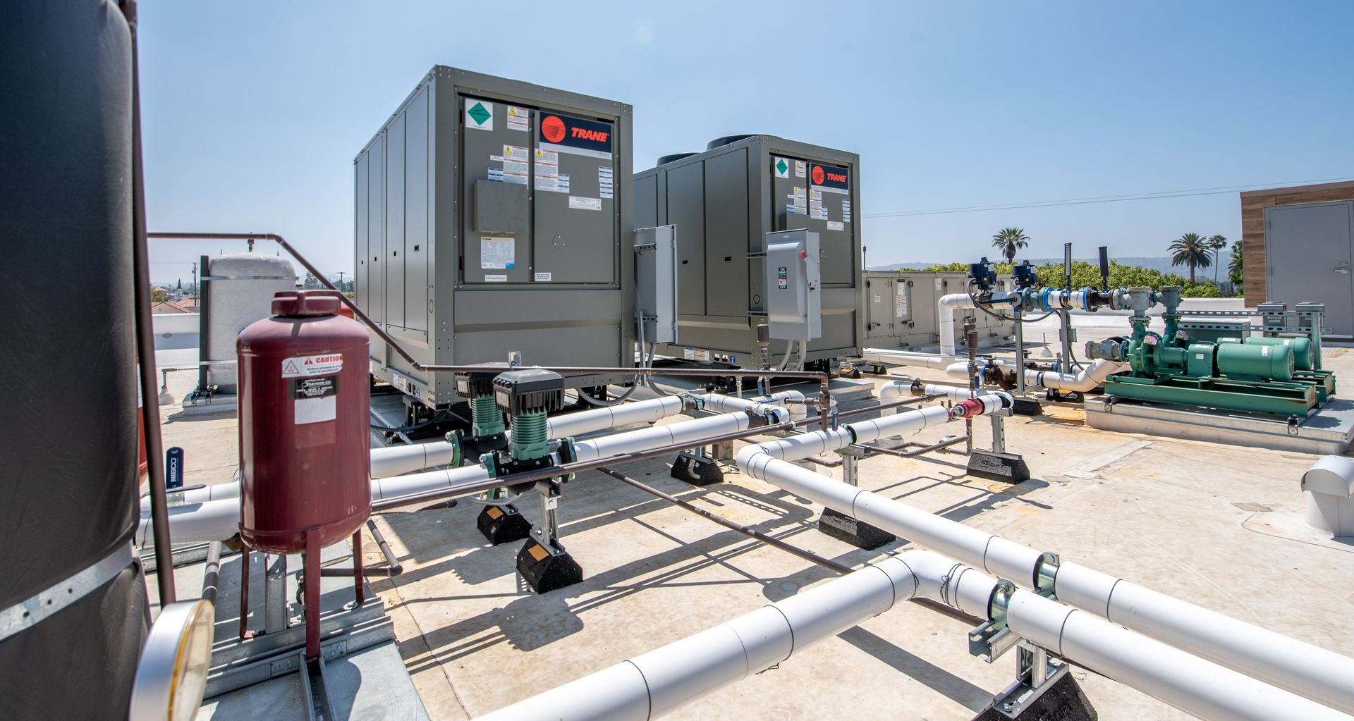 Rooftop HVAC units with pipes and a red tank, set against a bright blue sky.