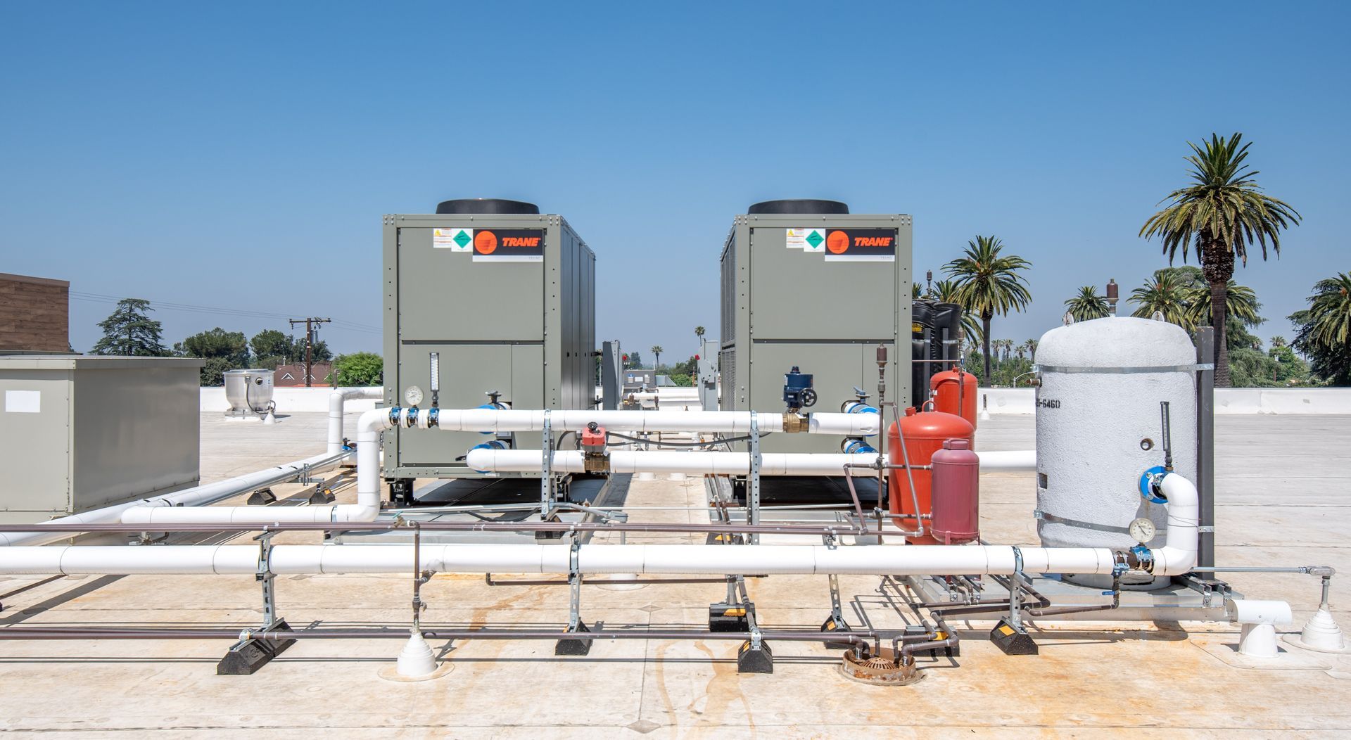 Rooftop HVAC units with pipes, valves, and a tank under a clear blue sky.