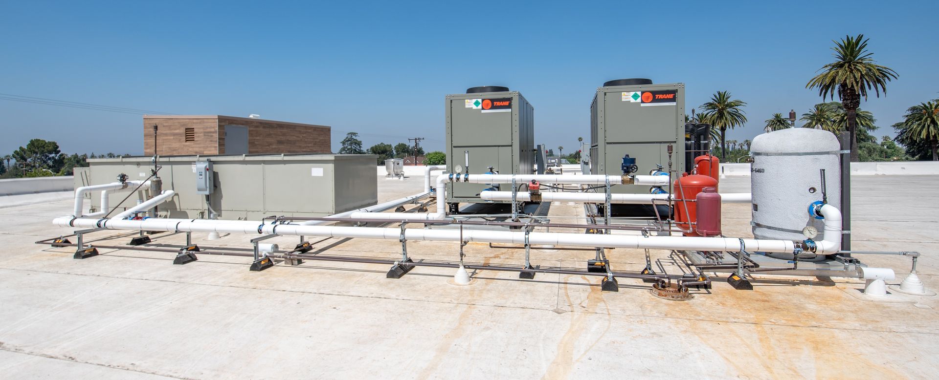 HVAC units and associated piping on a rooftop under a blue sky with palm trees in the distance.
