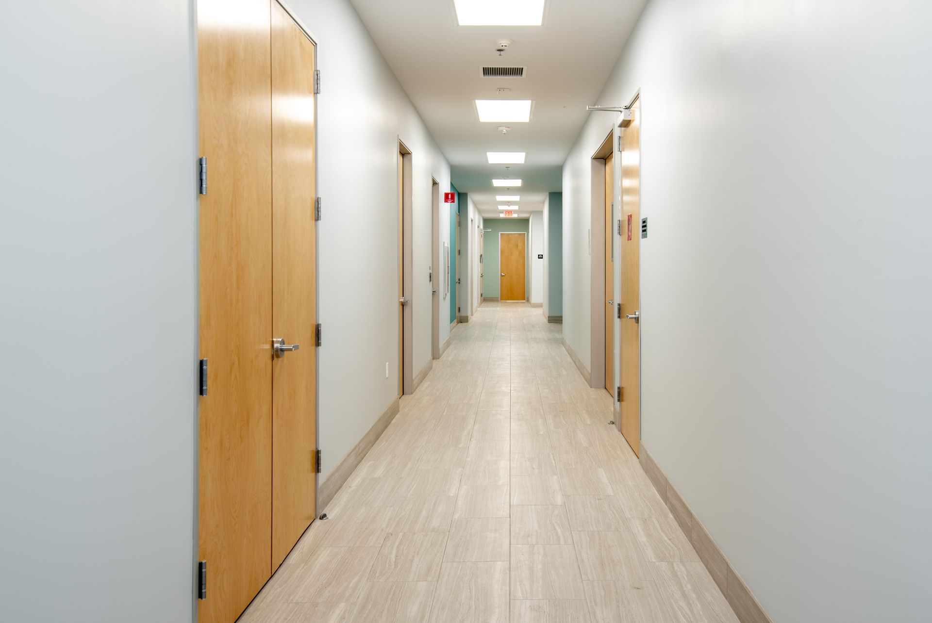 Long hallway with light blue walls, beige flooring, and wooden doors on either side.