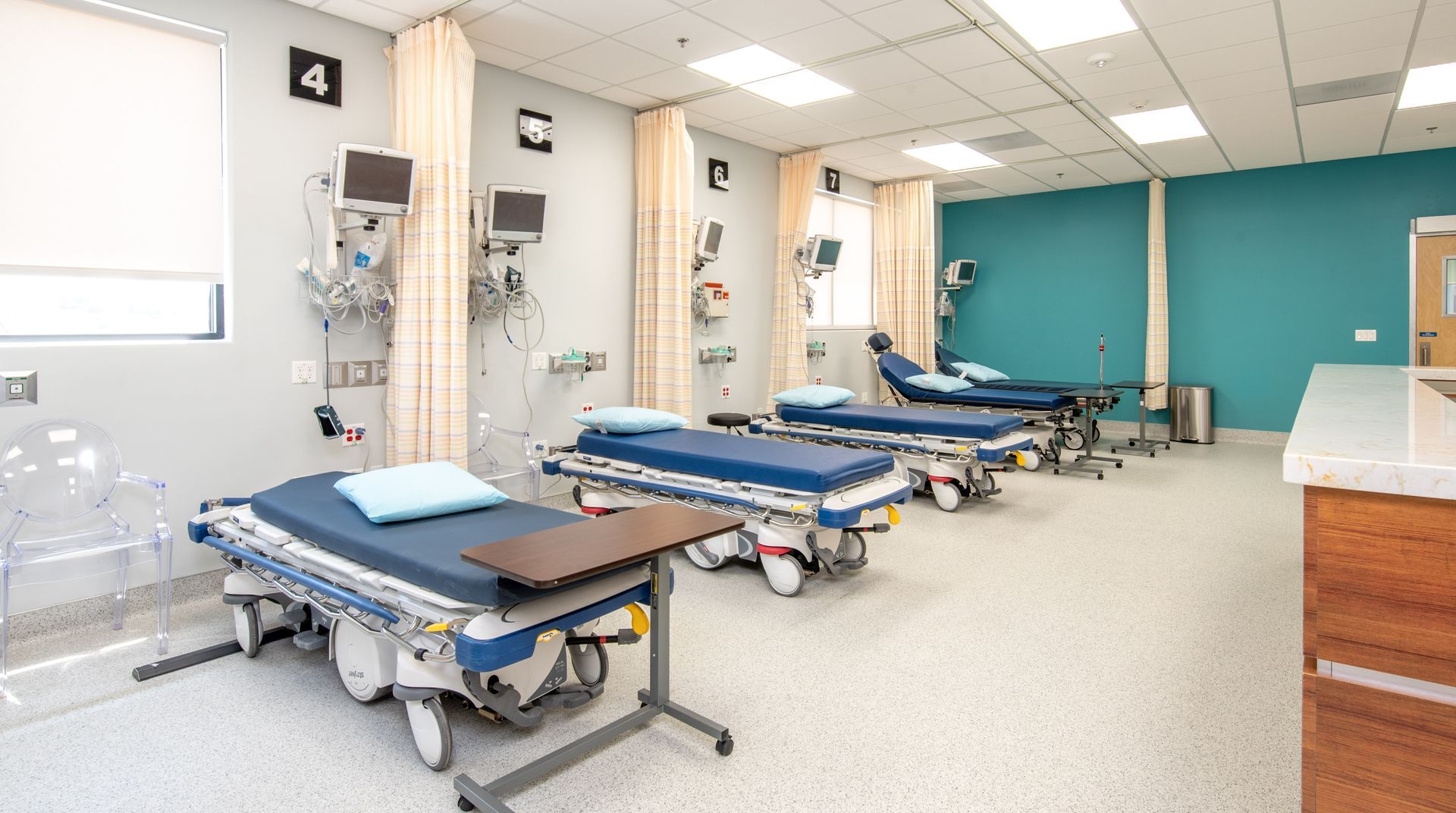 Emergency room with four beds, medical equipment, curtains, and teal and gray walls.