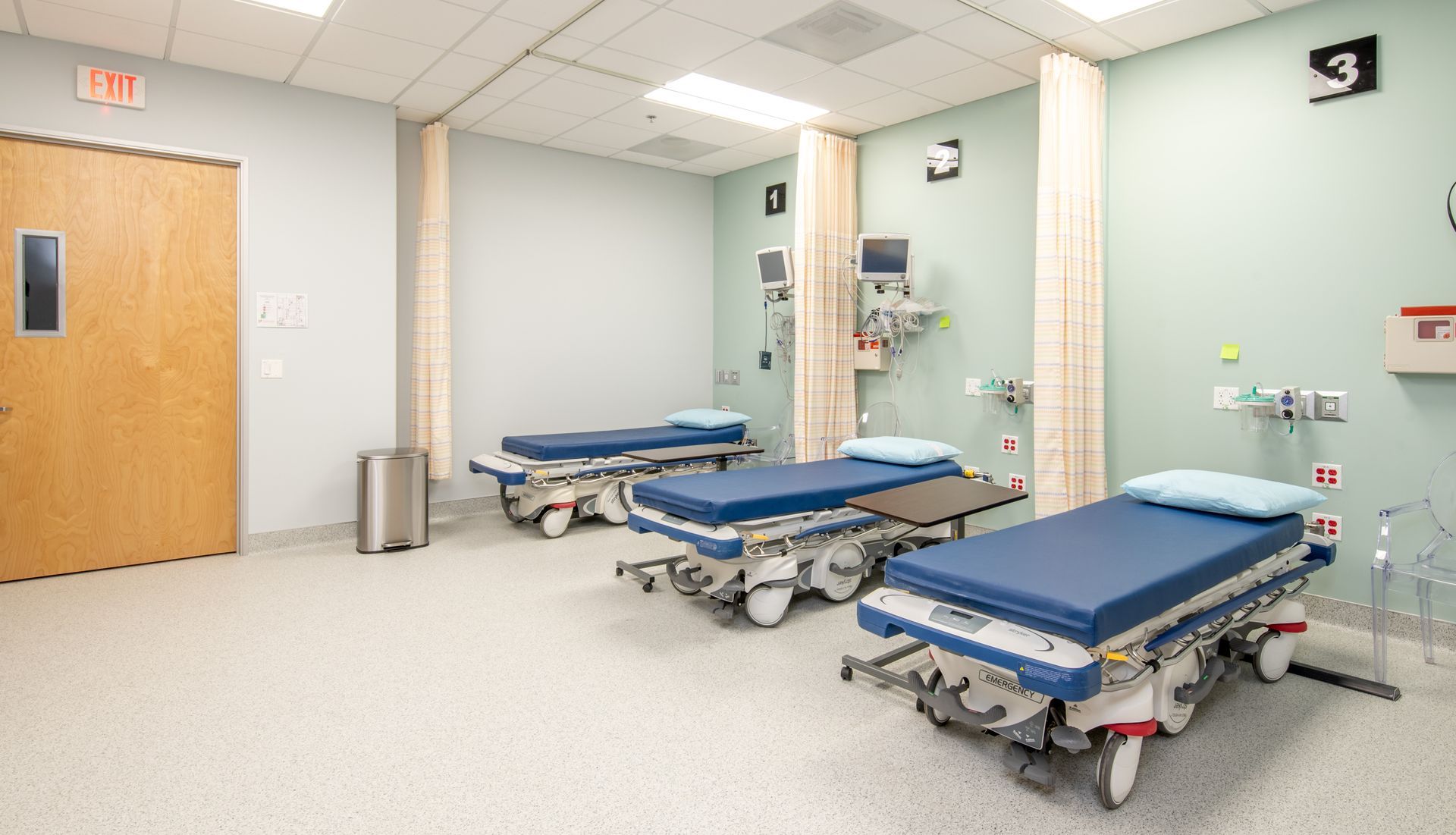 Three hospital beds in a light blue room with medical equipment; emergency room setting.