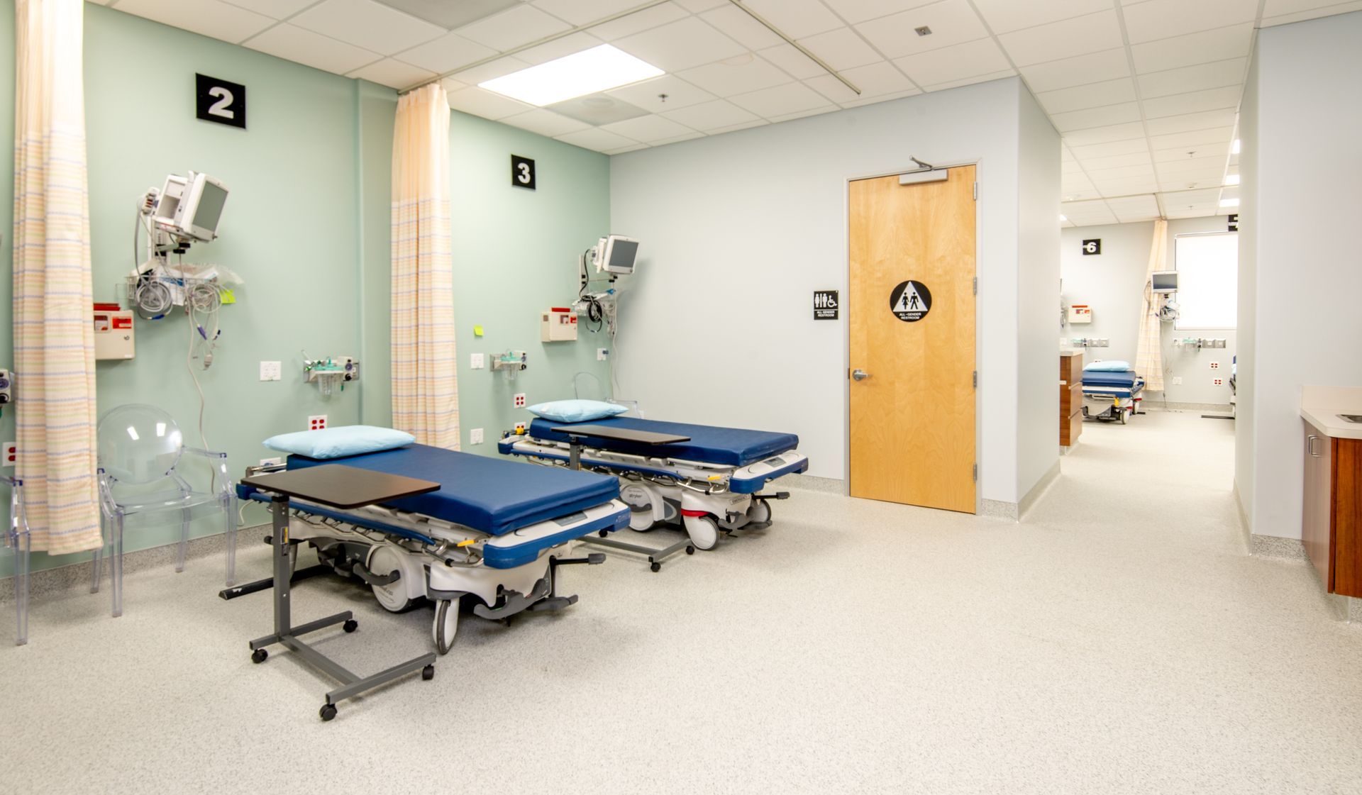Two hospital beds in a medical room with medical equipment. Beige and blue tones, pale green walls.