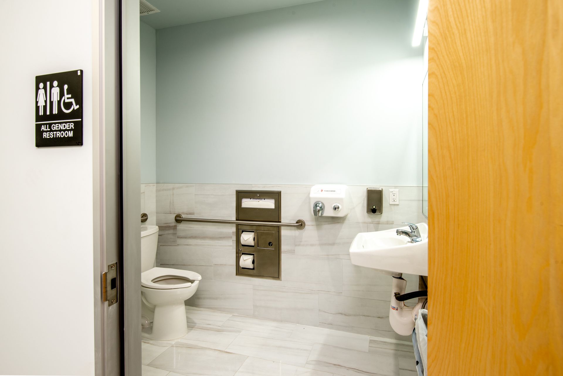 An accessible public restroom with a toilet, sink, and grab bars; a sign indicates it's inclusive.