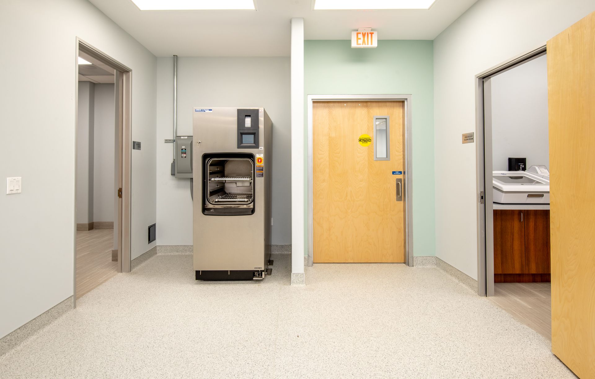 A sterilization room with a large machine. Doors on either side of a light-colored room with white speckled flooring.