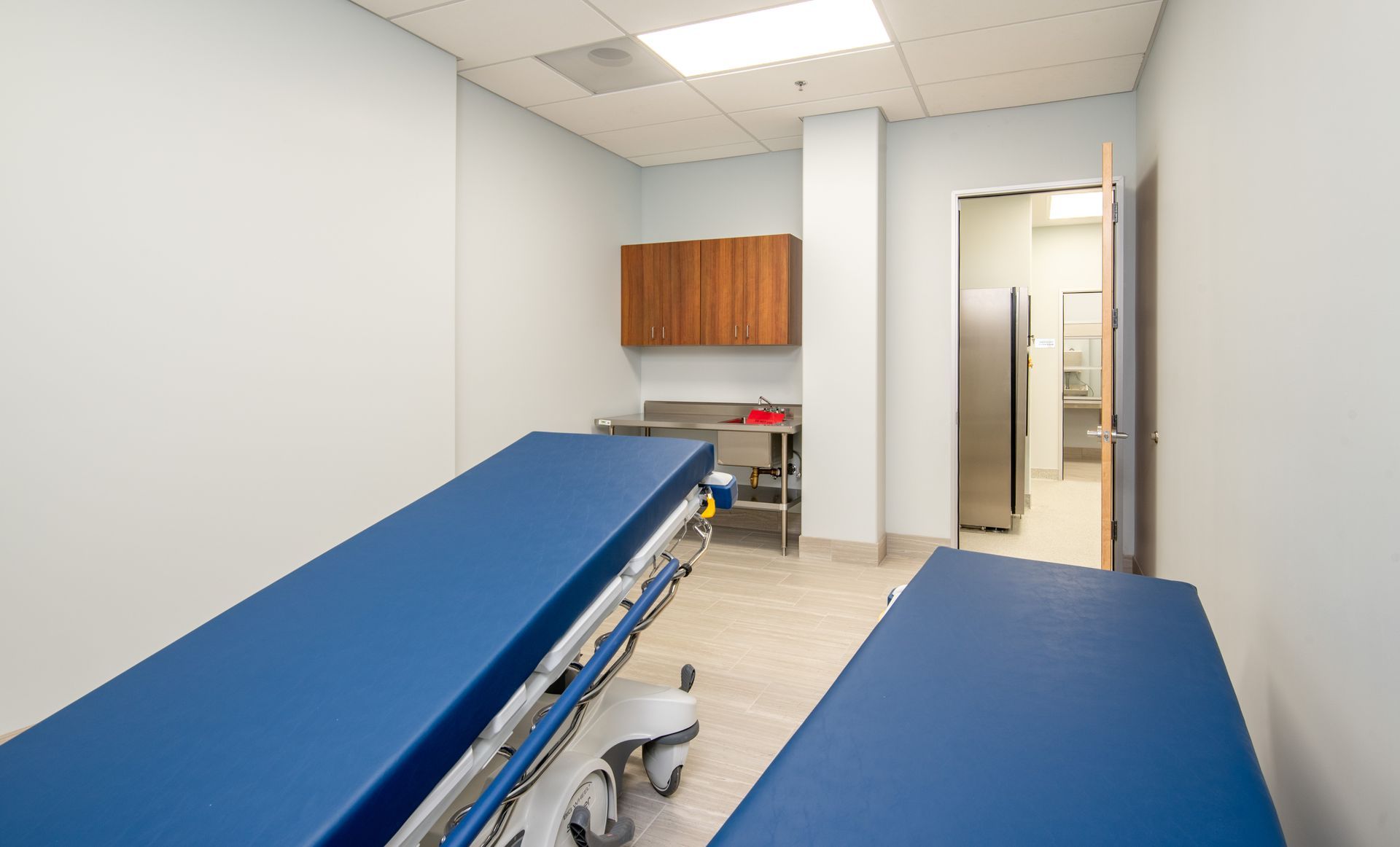 Medical exam room with two blue-topped beds, cabinets, and an open doorway to a bathroom.
