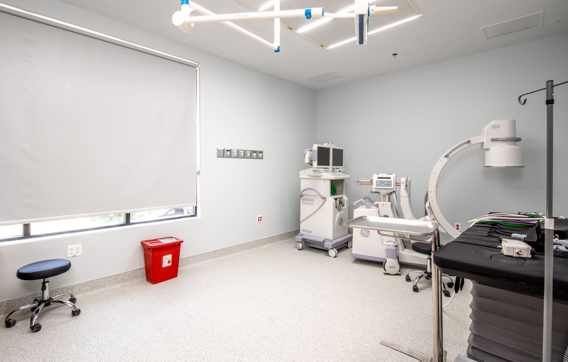 Medical room with equipment: light-colored walls, surgical table, monitoring devices, and a window.