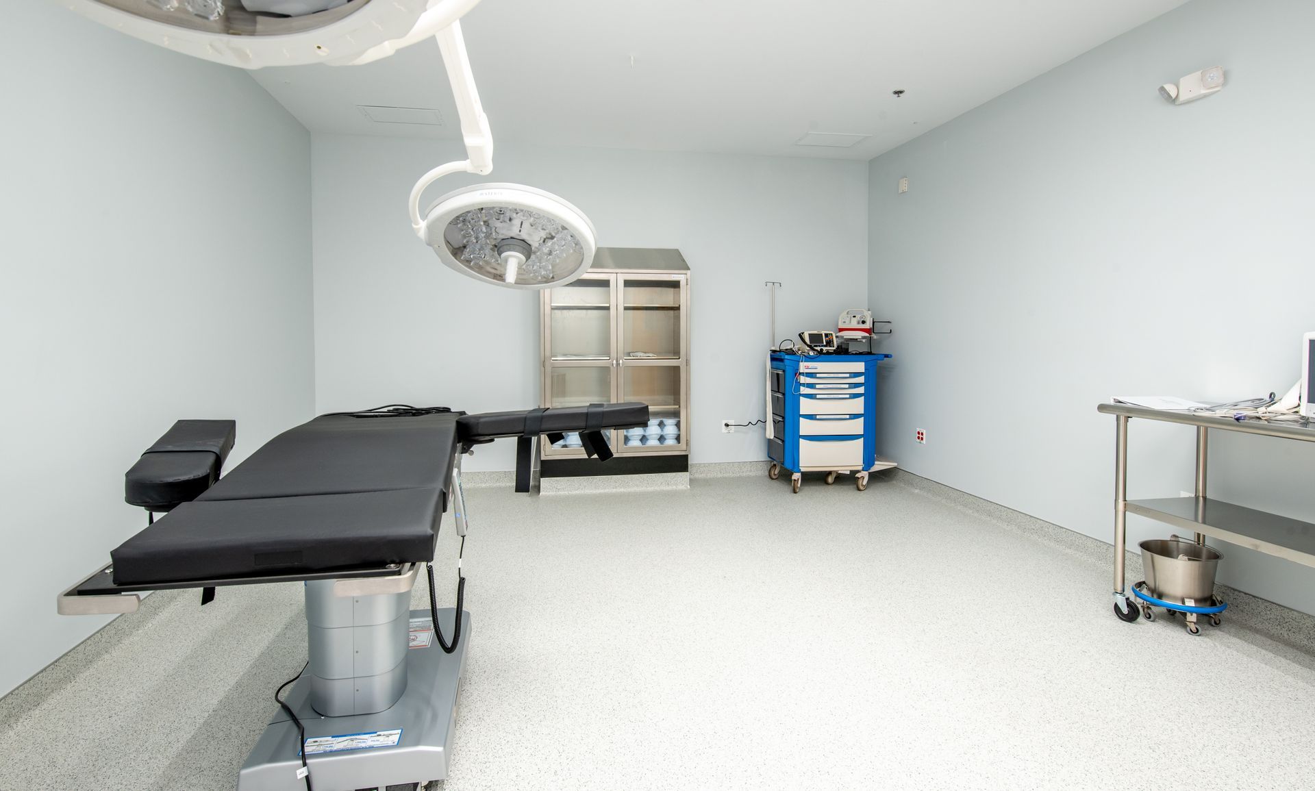 Operating room with surgical bed, overhead lights, medical equipment, and pale walls.