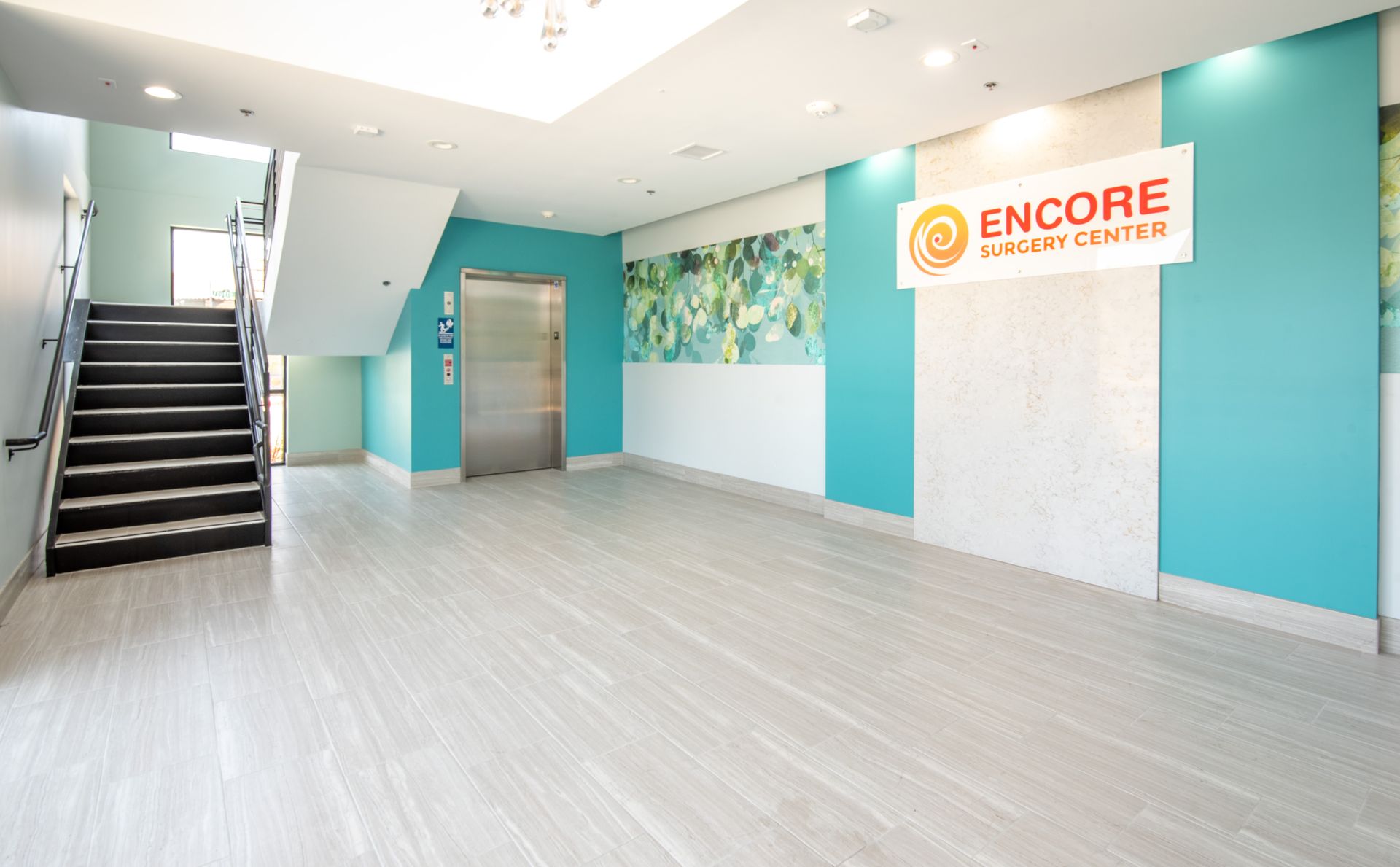 Lobby of Encore Surgery Center. Aqua and white walls, gray stairs, and a silver elevator. The logo is visible.