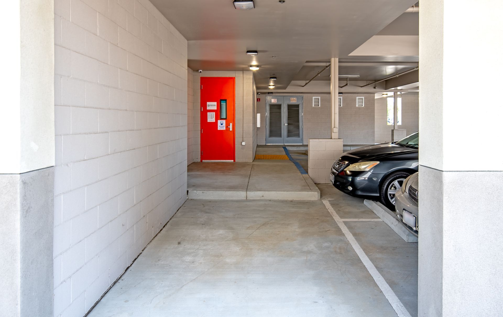 Concrete corridor with a red door, leading to a parking area with a car.