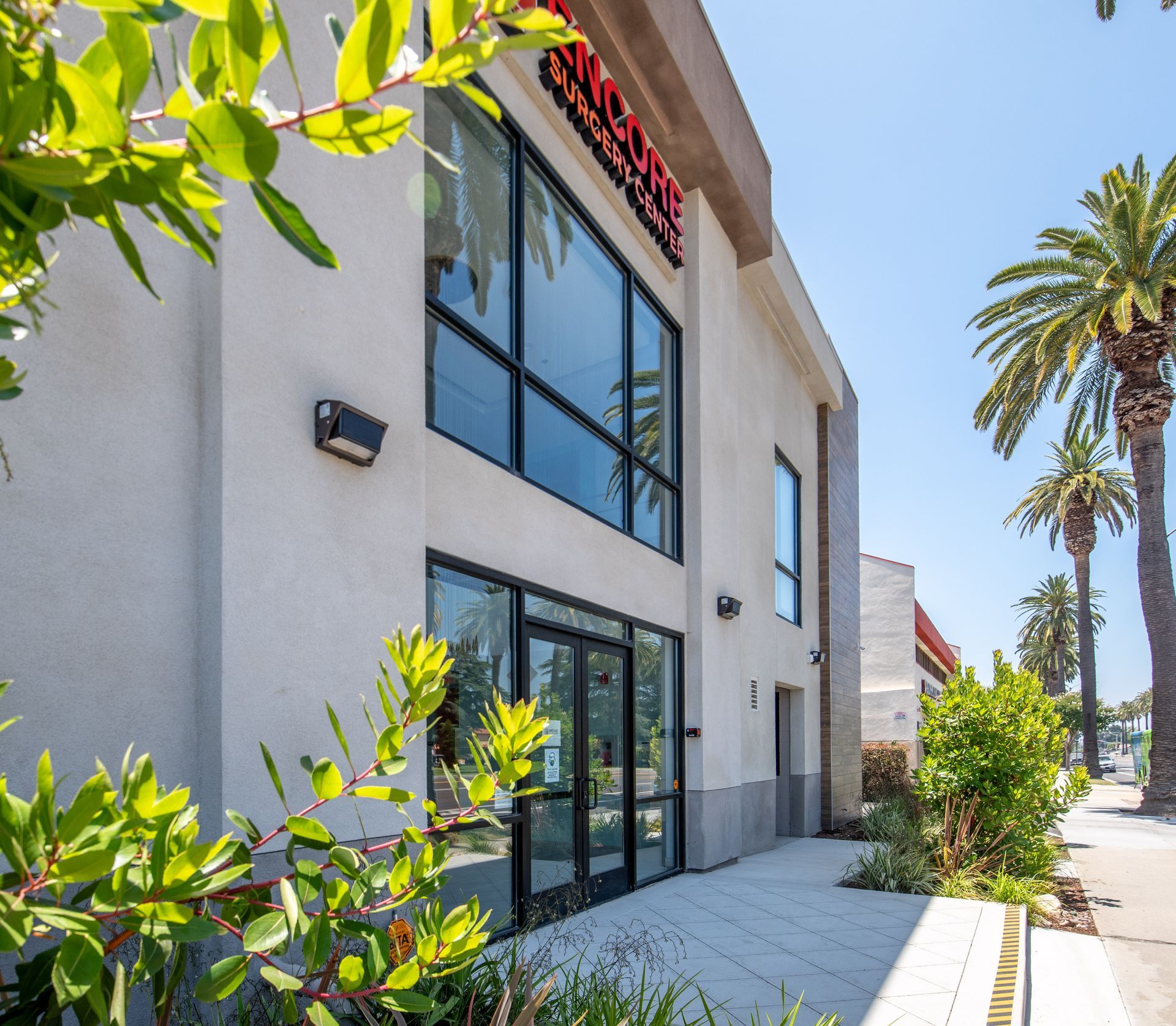 Exterior view of a building with large windows, a walkway, and palm trees, with bright sunshine.