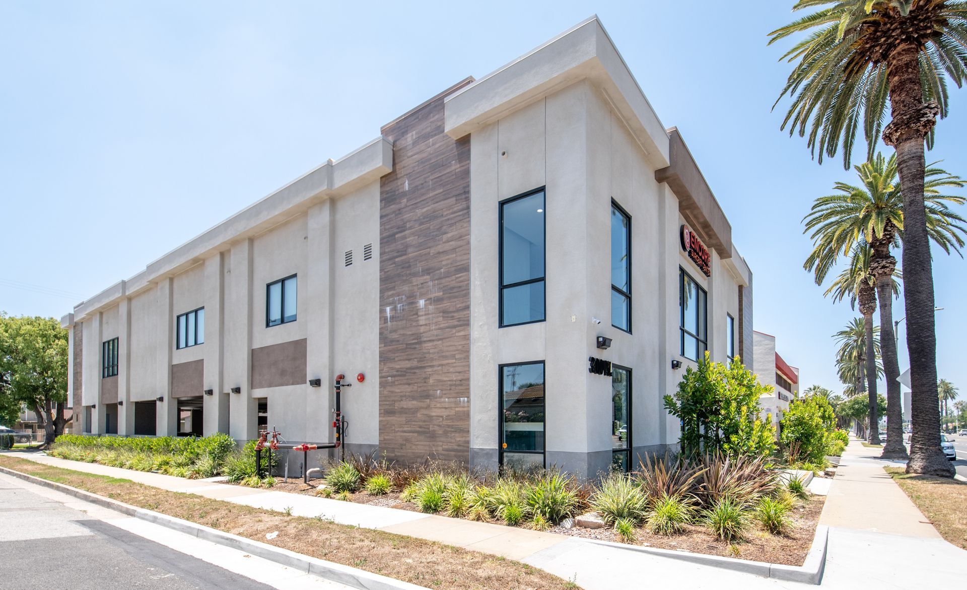 Two-story commercial building with beige and brick facade, large windows, and palm trees on the sidewalk.