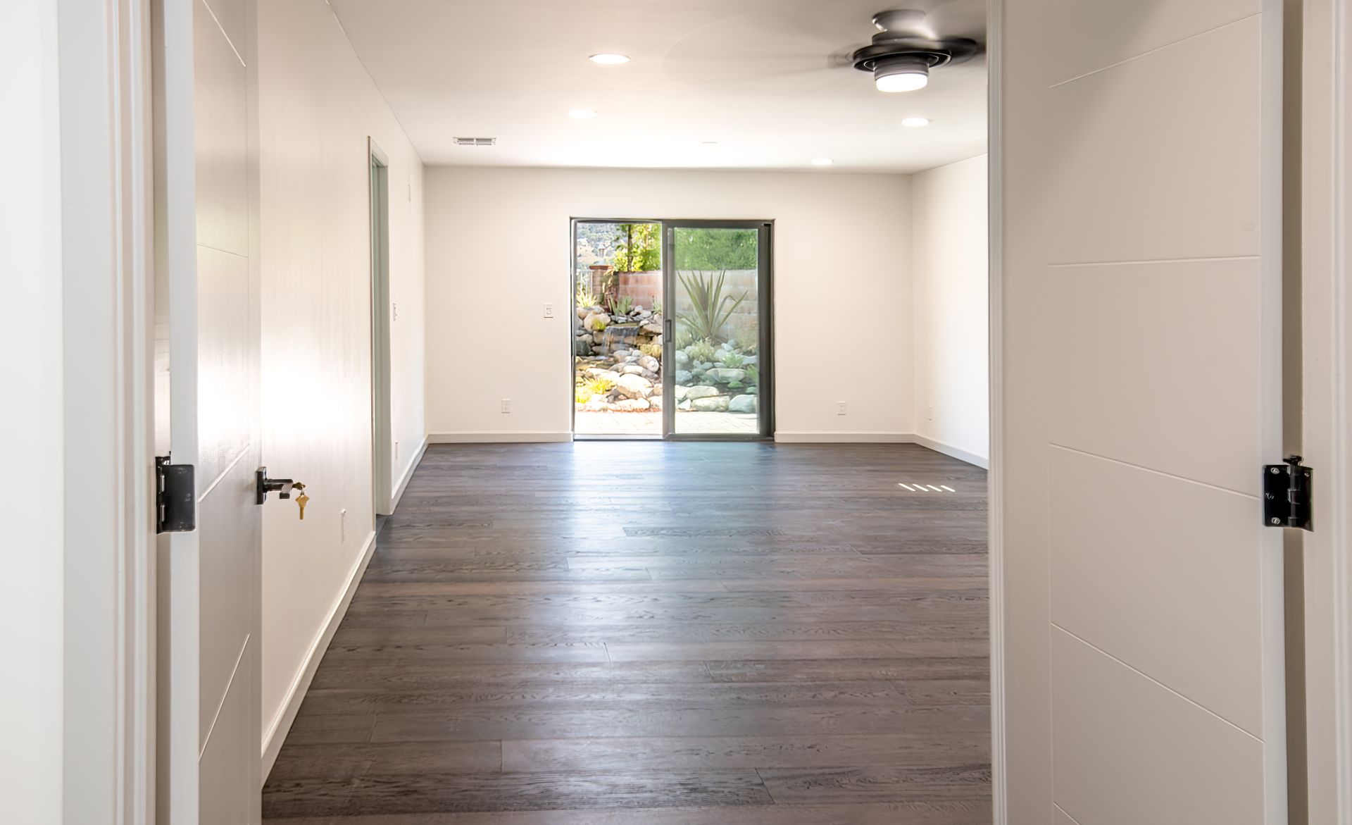 Empty room with dark wood floors, white walls, and sliding glass doors to a garden.