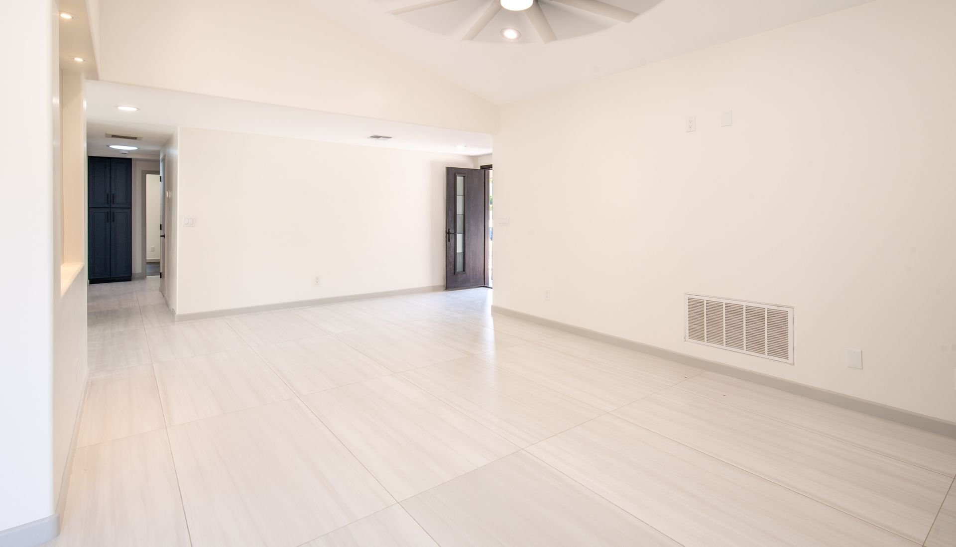 Empty, bright living room with white tile floor and walls; a dark door is visible.