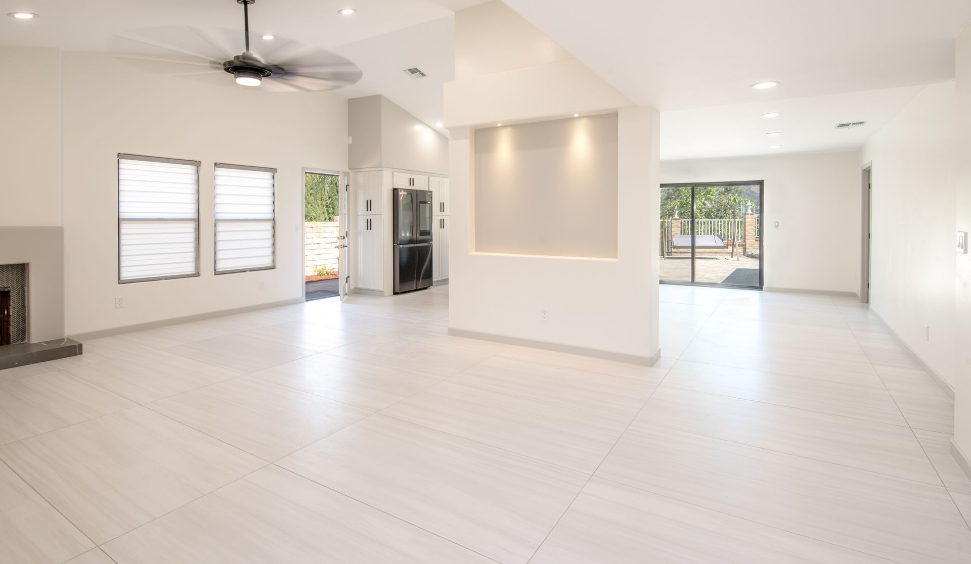 Empty, bright white living room with tiled floor, three windows, and a ceiling fan.