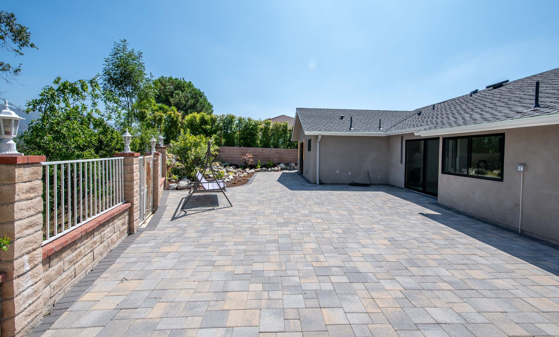 Backyard patio with brick pavers, partial fence, and house under blue sky.