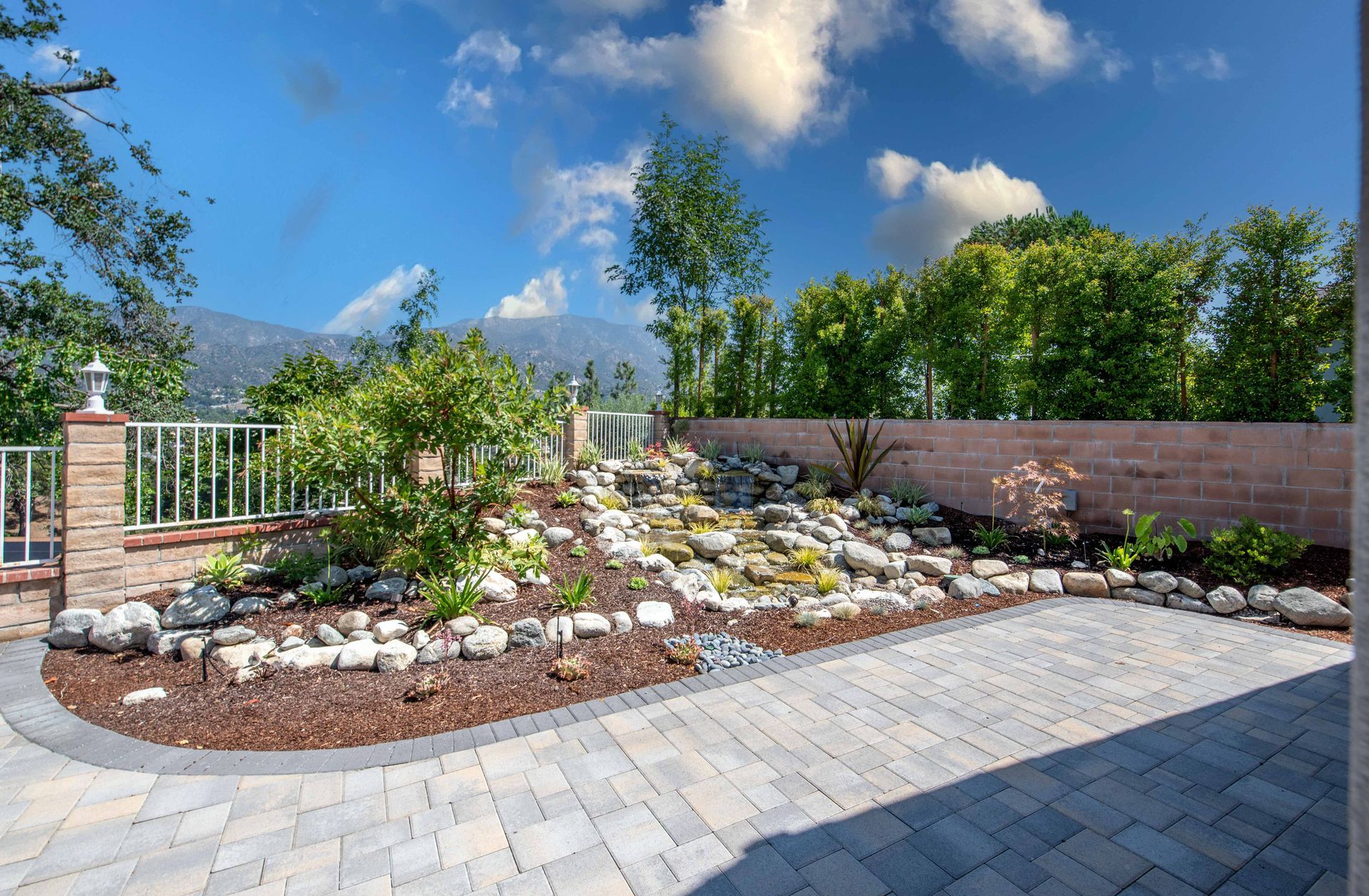 Landscaped yard with rock garden, patio, trees, fence, mountains, and blue sky.