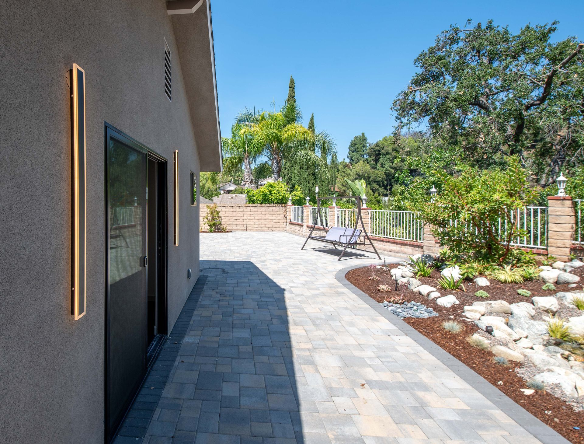 Exterior view with stone pathway leading to a backyard with swing, greenery, and a blue sky.