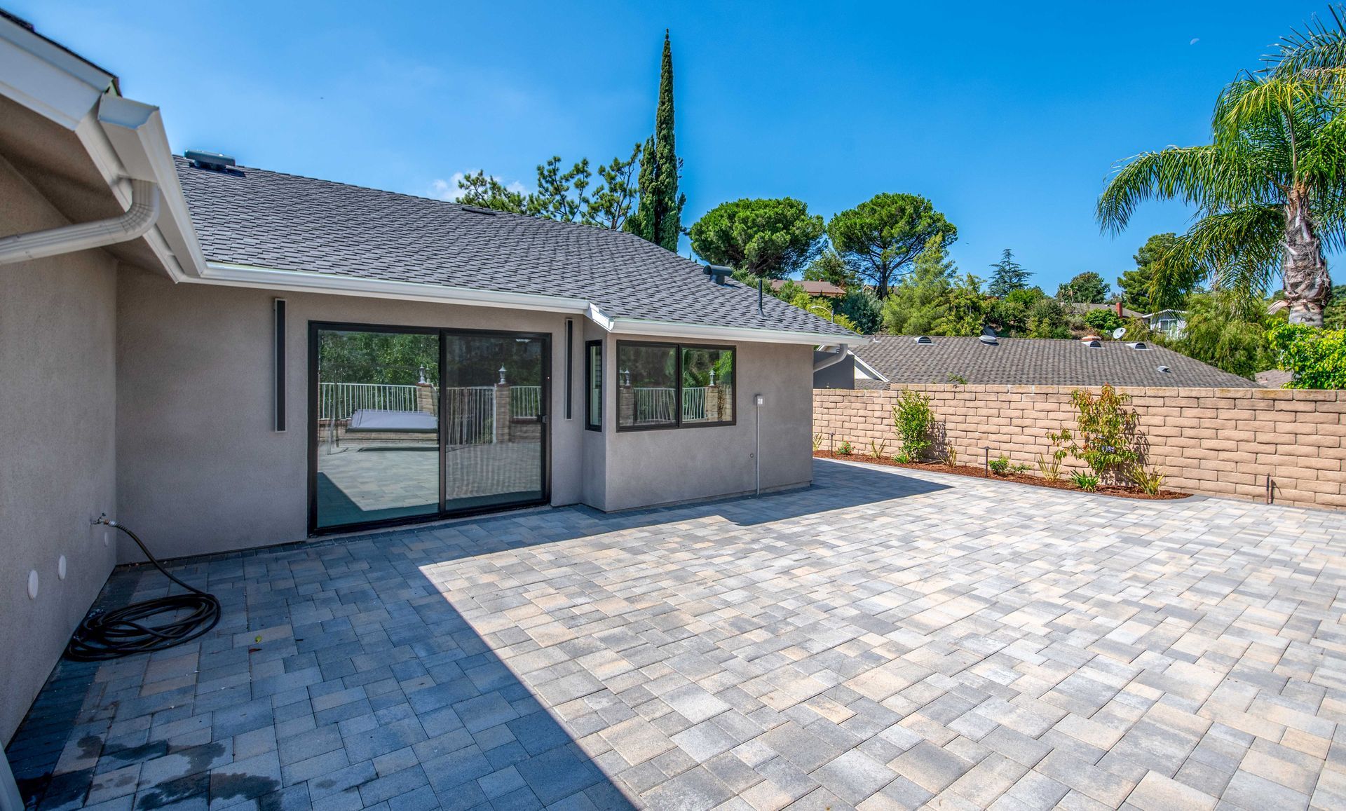 Backyard patio with stone pavers, sliding glass door, and brick wall under a blue sky.