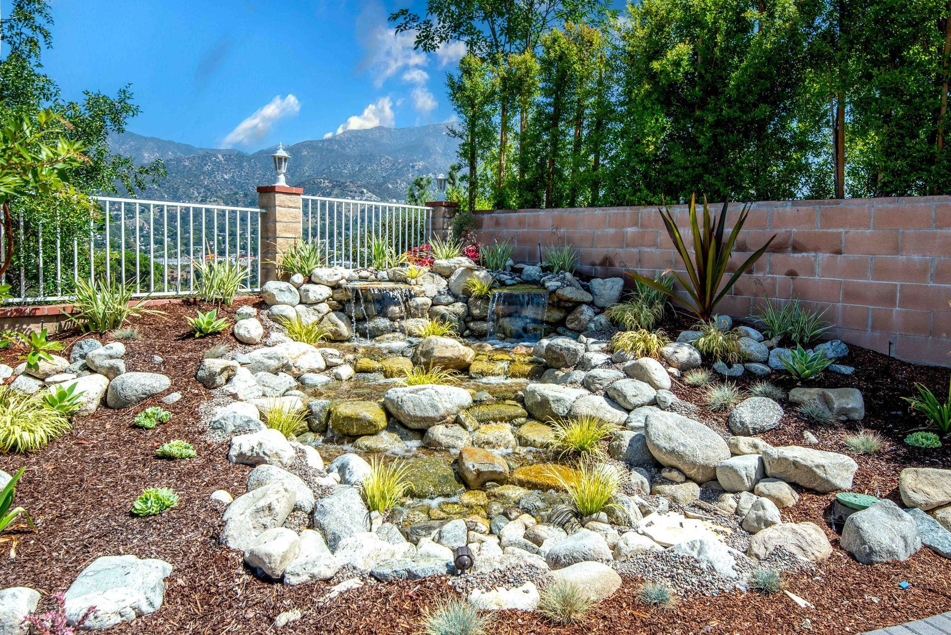 A cascading rock waterfall in a landscaped backyard with greenery and a brick wall.