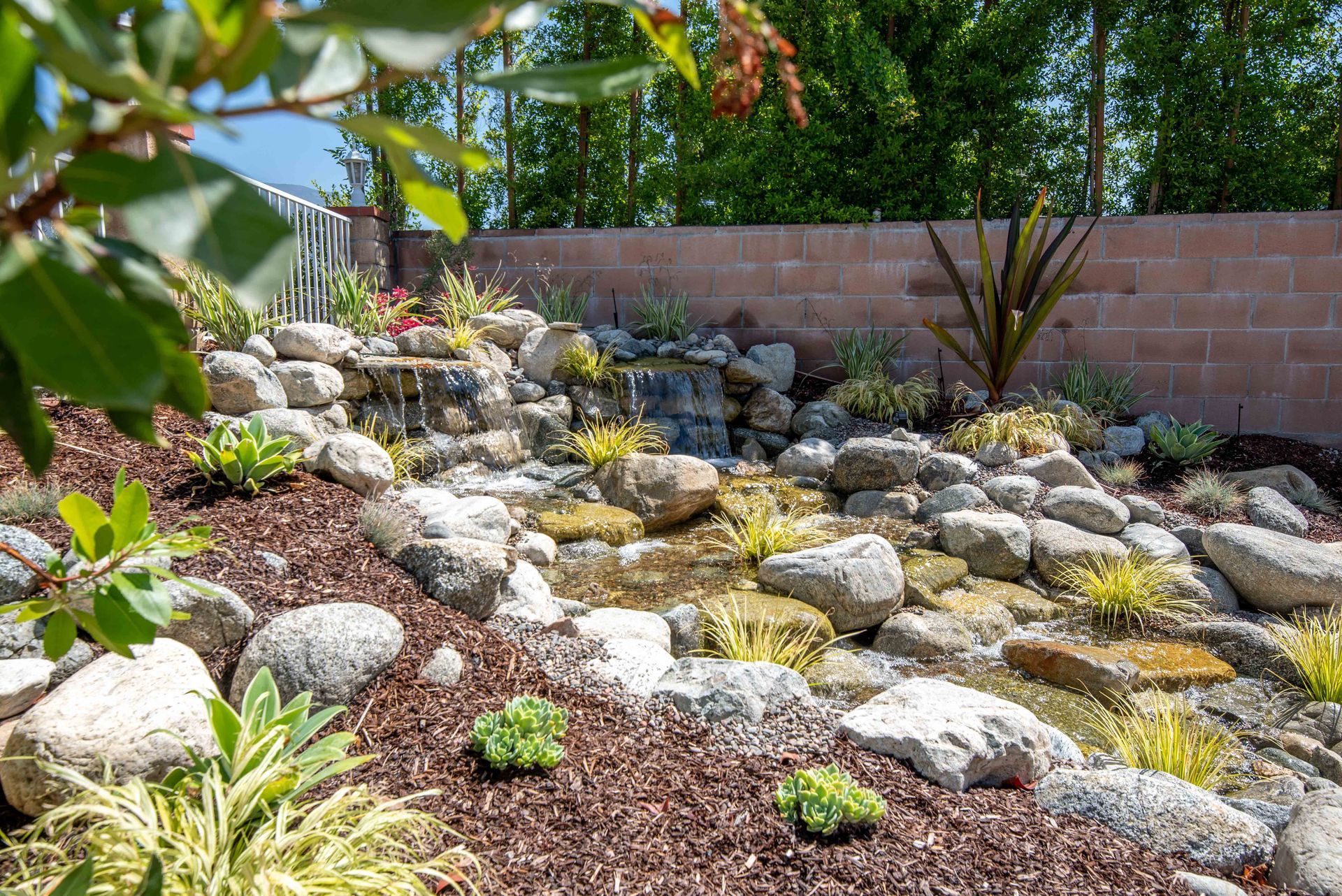 Water feature cascading over rocks in a landscaped garden, brown mulch, green and yellow plants.
