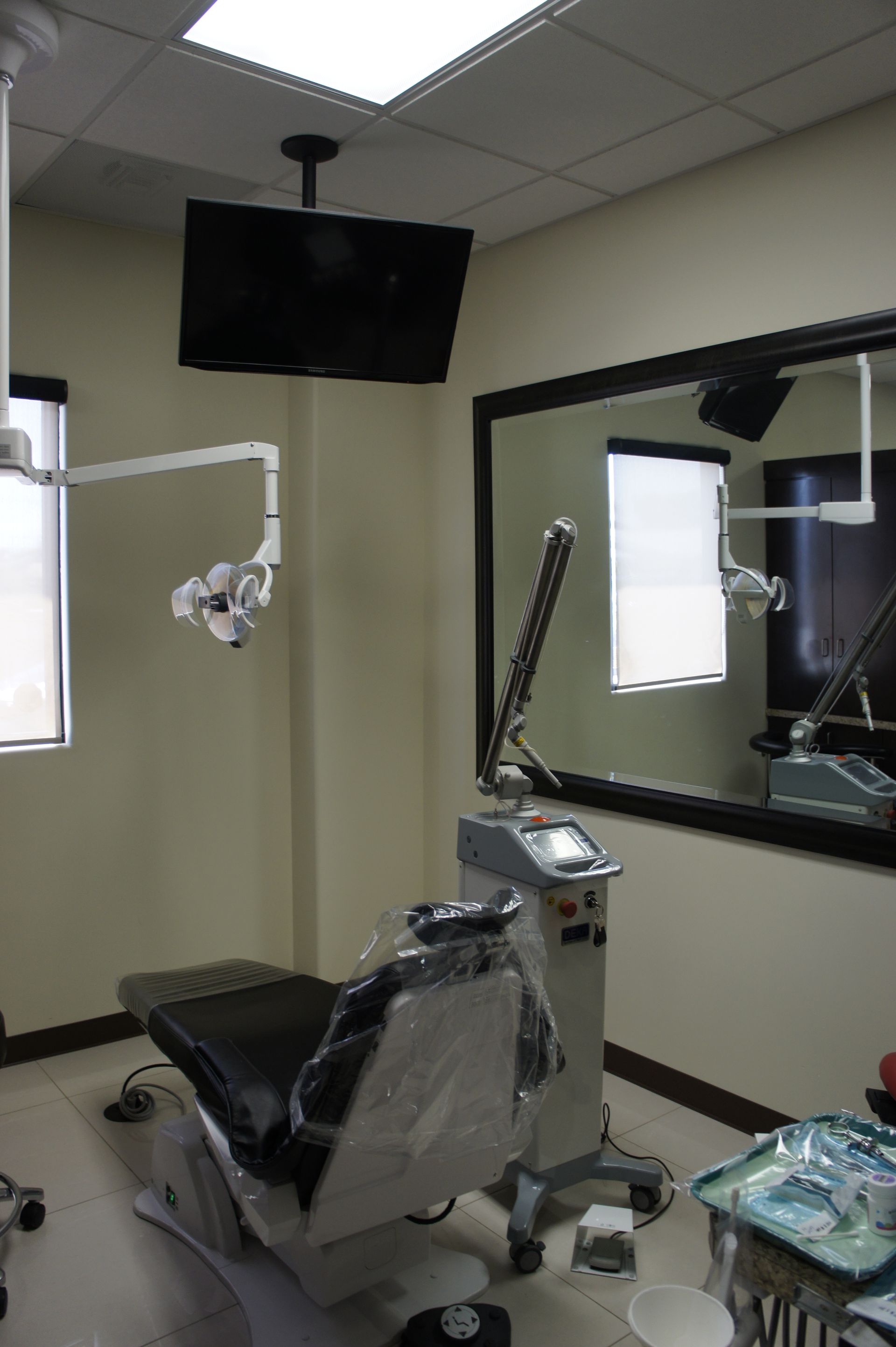 Dental office interior: dental chair, equipment, TV, large mirror reflecting another room.