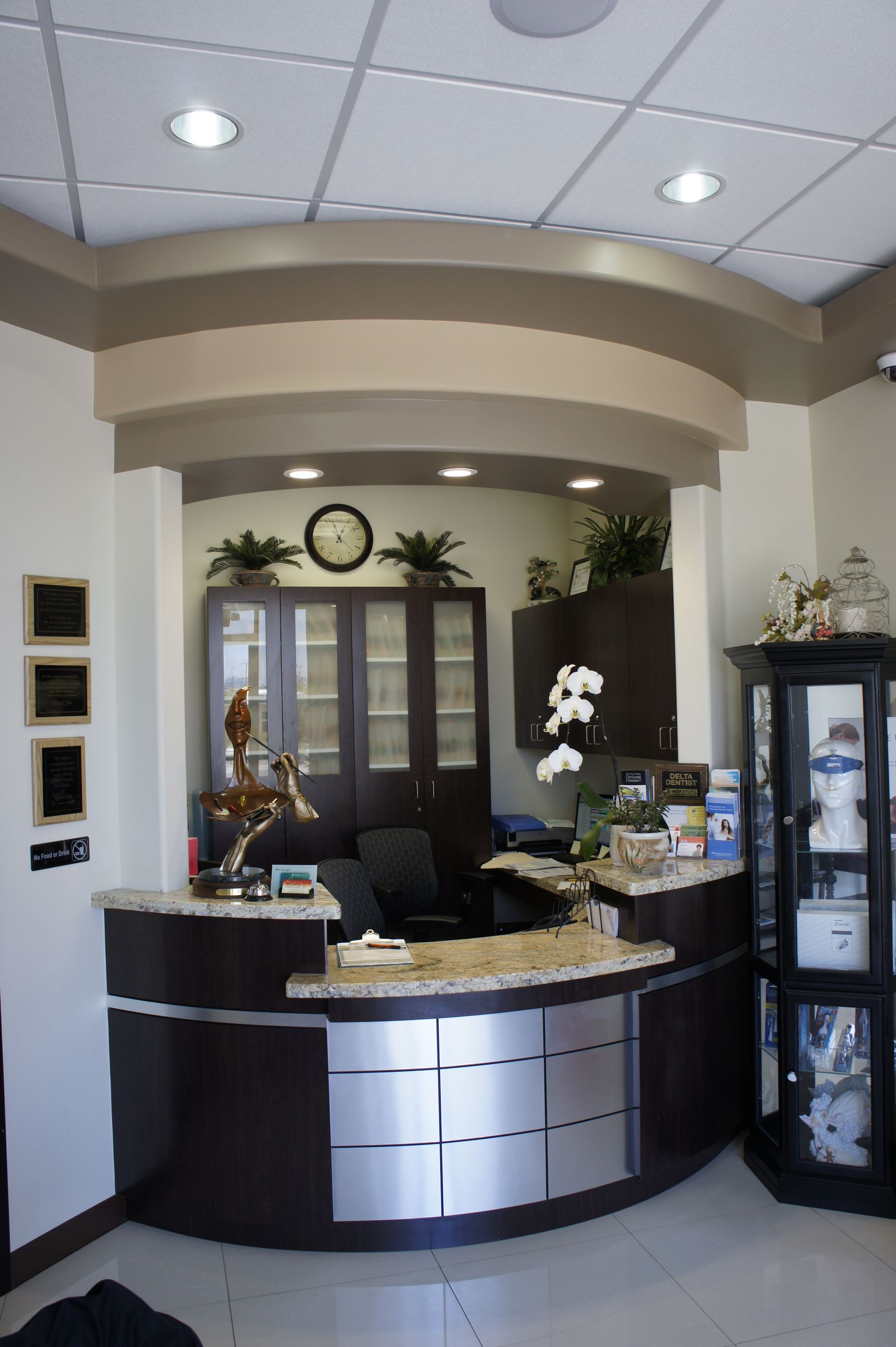 Reception desk in a dental office. Dark wood, stainless steel, plants, clock, and framed items.