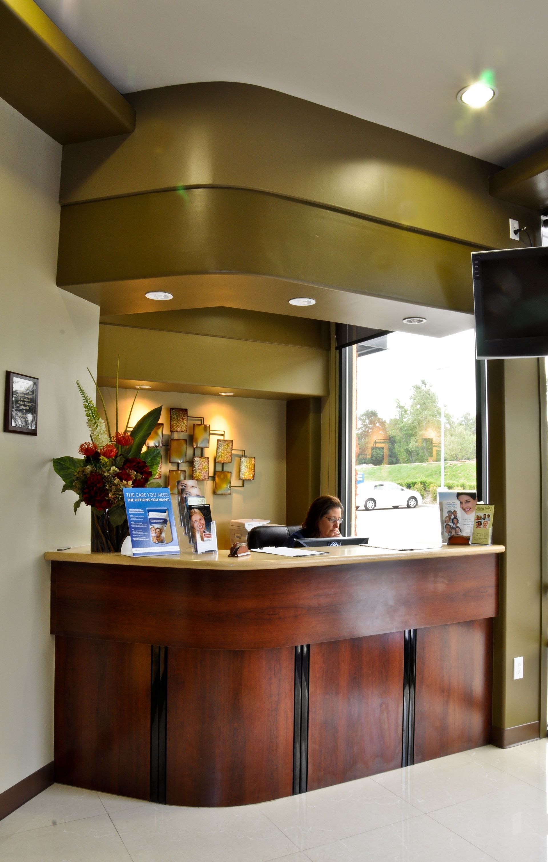 Reception desk in a dental office; dark wood, curved front; person at desk, gold ceiling, large window.