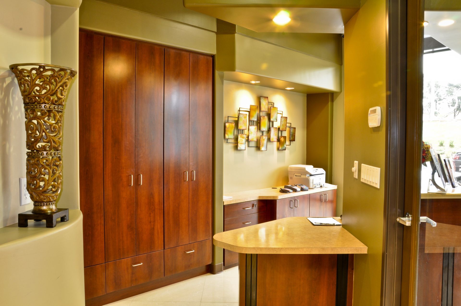 Reception area with wood cabinets, golden vase, and counter. Walls are olive green.