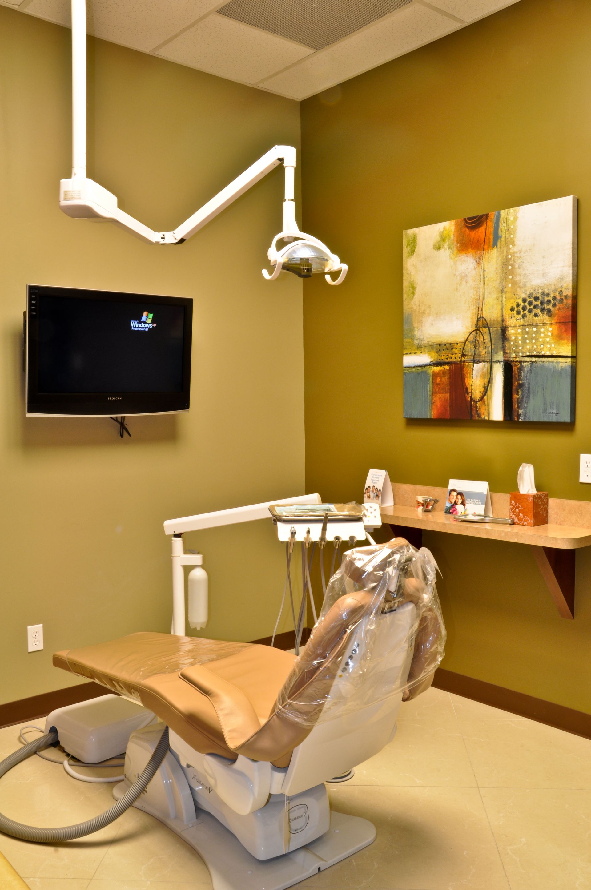 Dental exam room with chair, equipment, mounted TV, artwork, and overhead light.