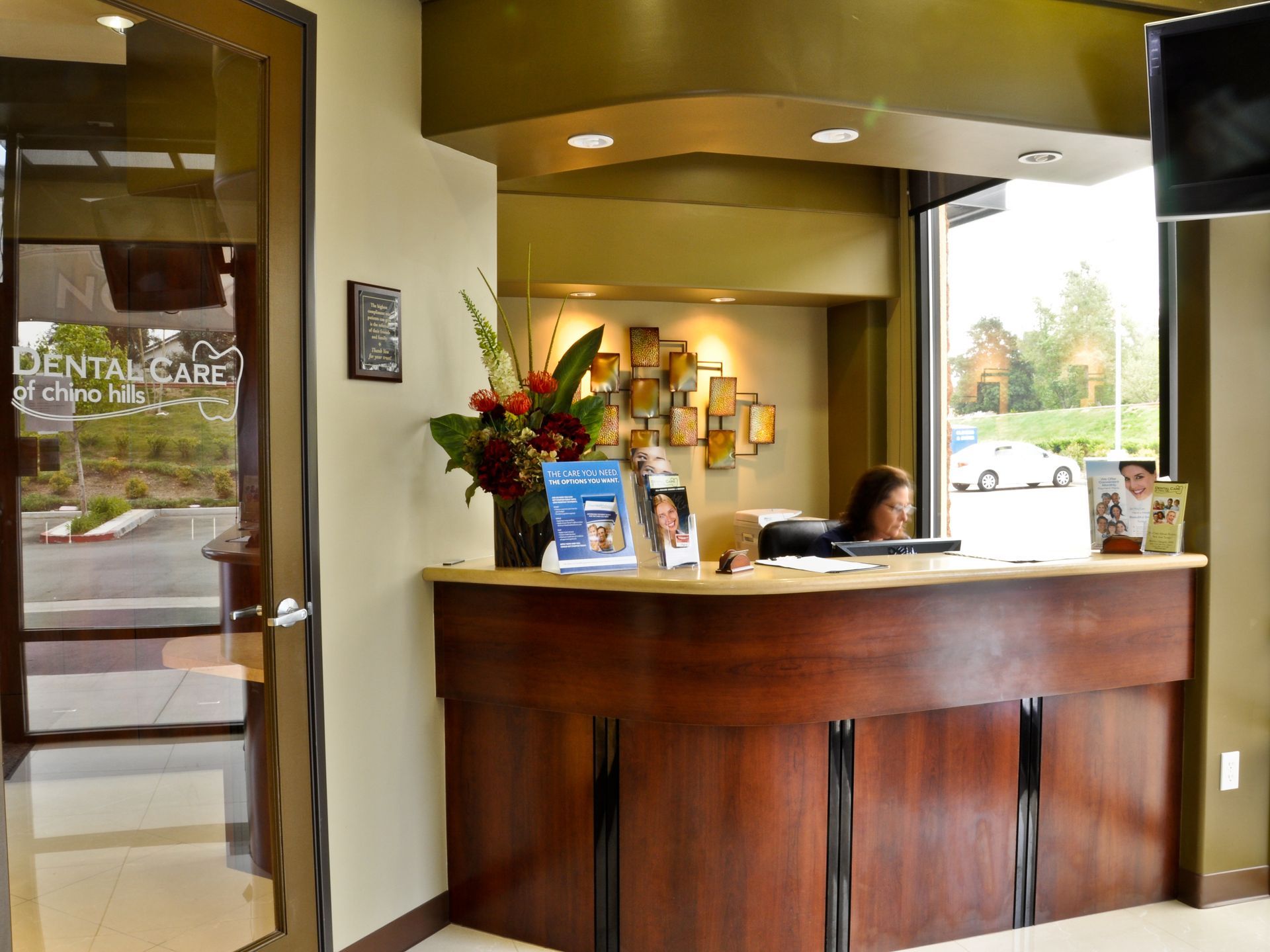Reception area of a dental clinic with a receptionist at the desk. Wooden finish, flowers, and advertisement display.
