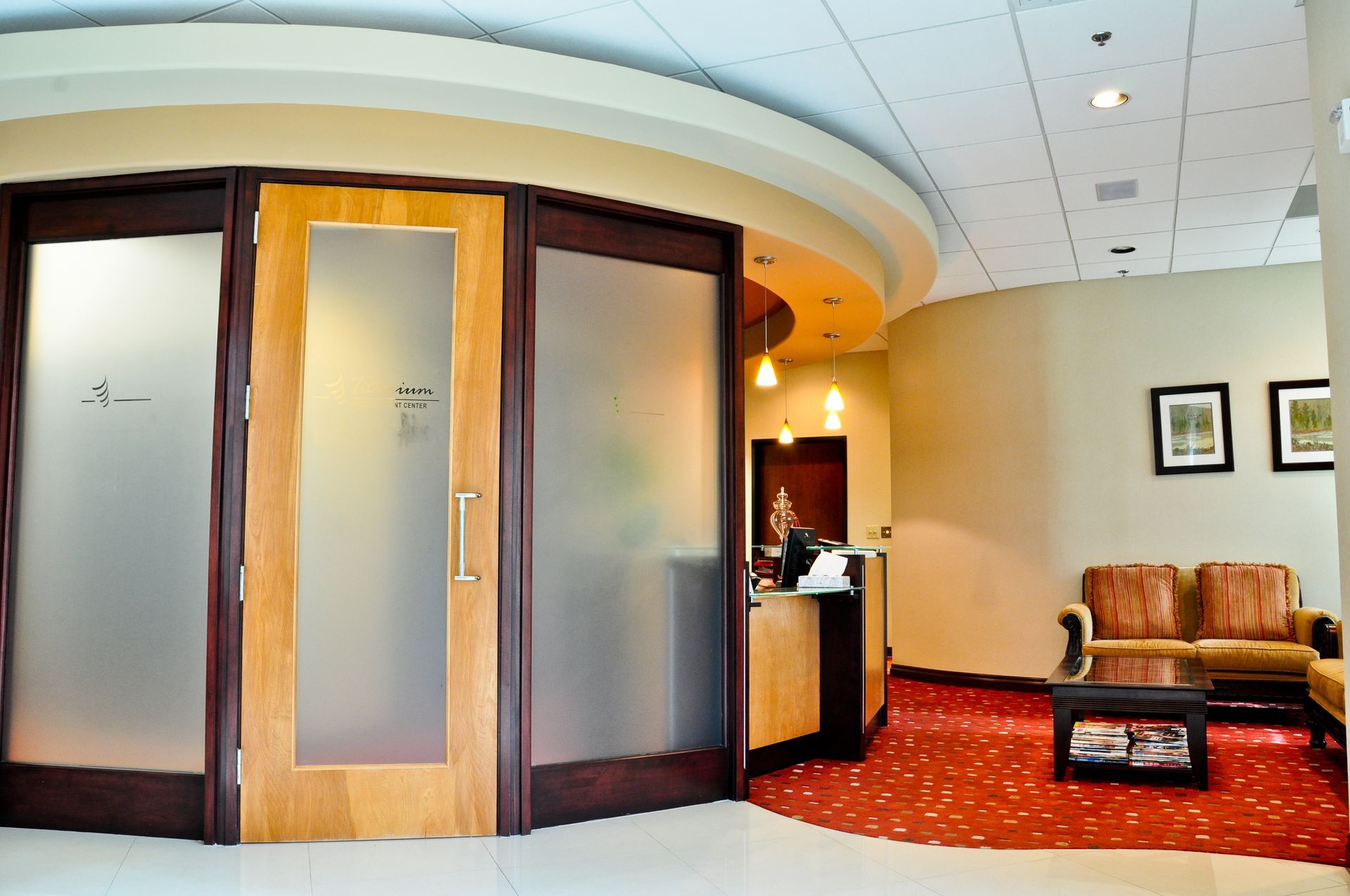Office reception area with frosted glass doors, a curved wall, red carpet, and a waiting area with a brown couch.