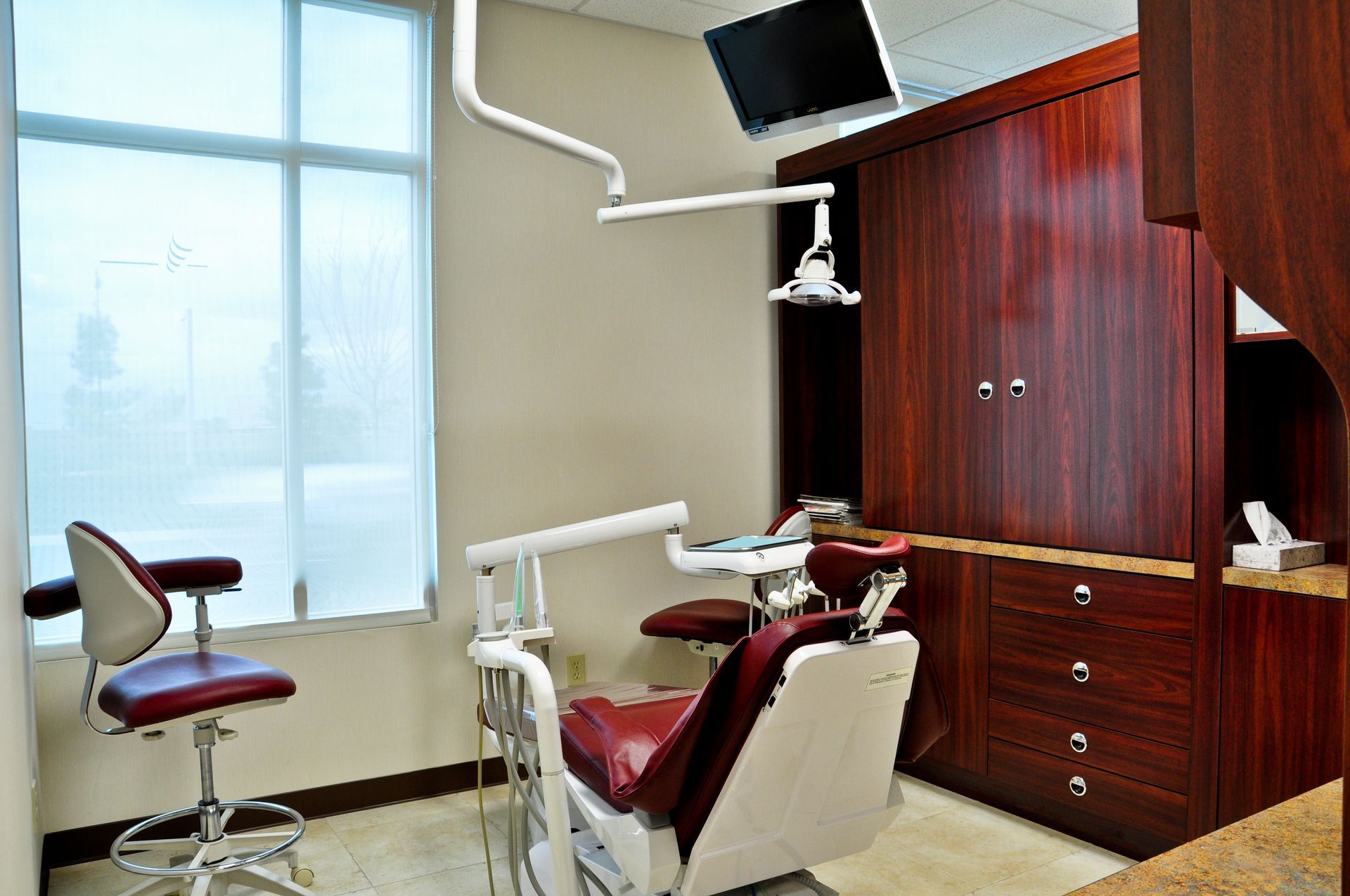 Dentist's office: red dental chair, window, brown cabinetry, overhead light, and small tv.