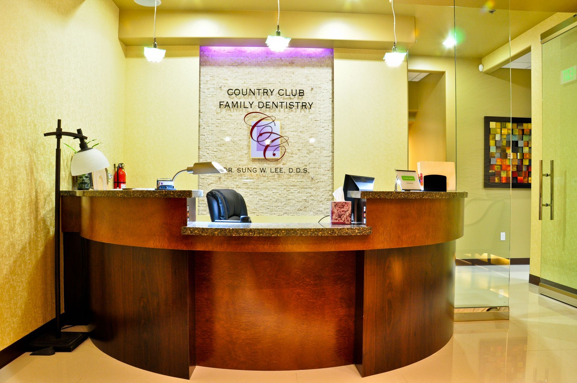 Reception desk in a dental office: brown wood, stone top, logo, cream walls, a painting and a frosted glass door.