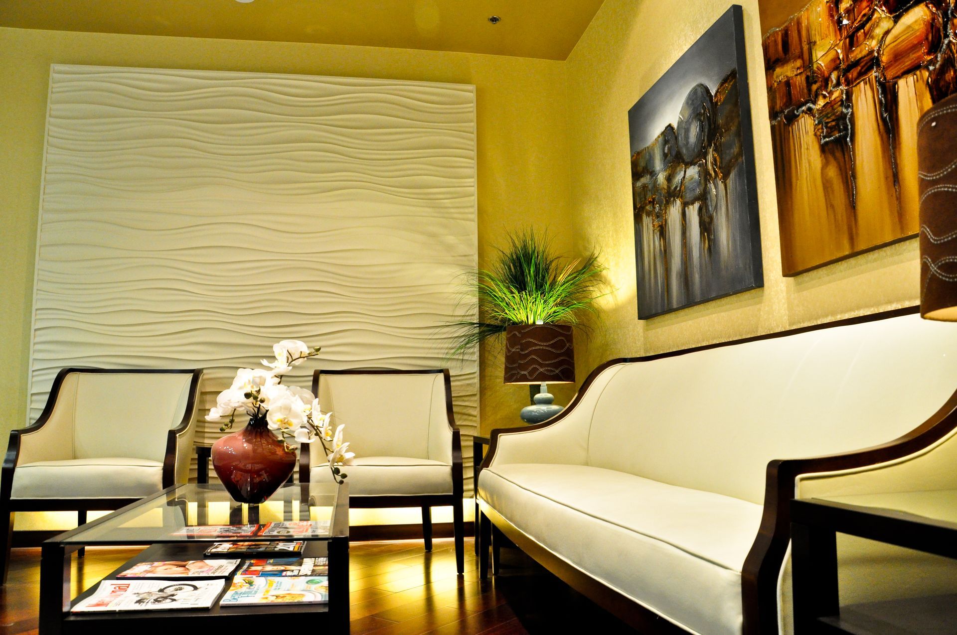 A waiting room with white furniture, art on walls, and a coffee table. Yellow walls, brown wooden floor.