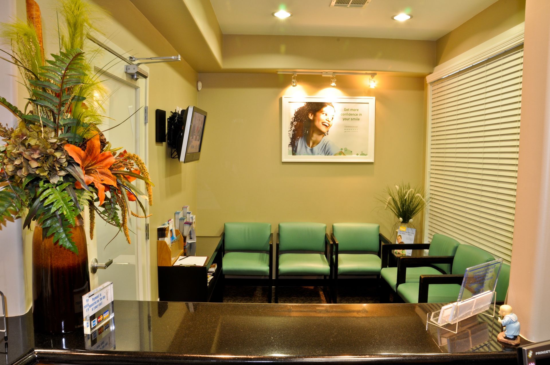 Dental office waiting room with green chairs, floral arrangement, and a reception desk.