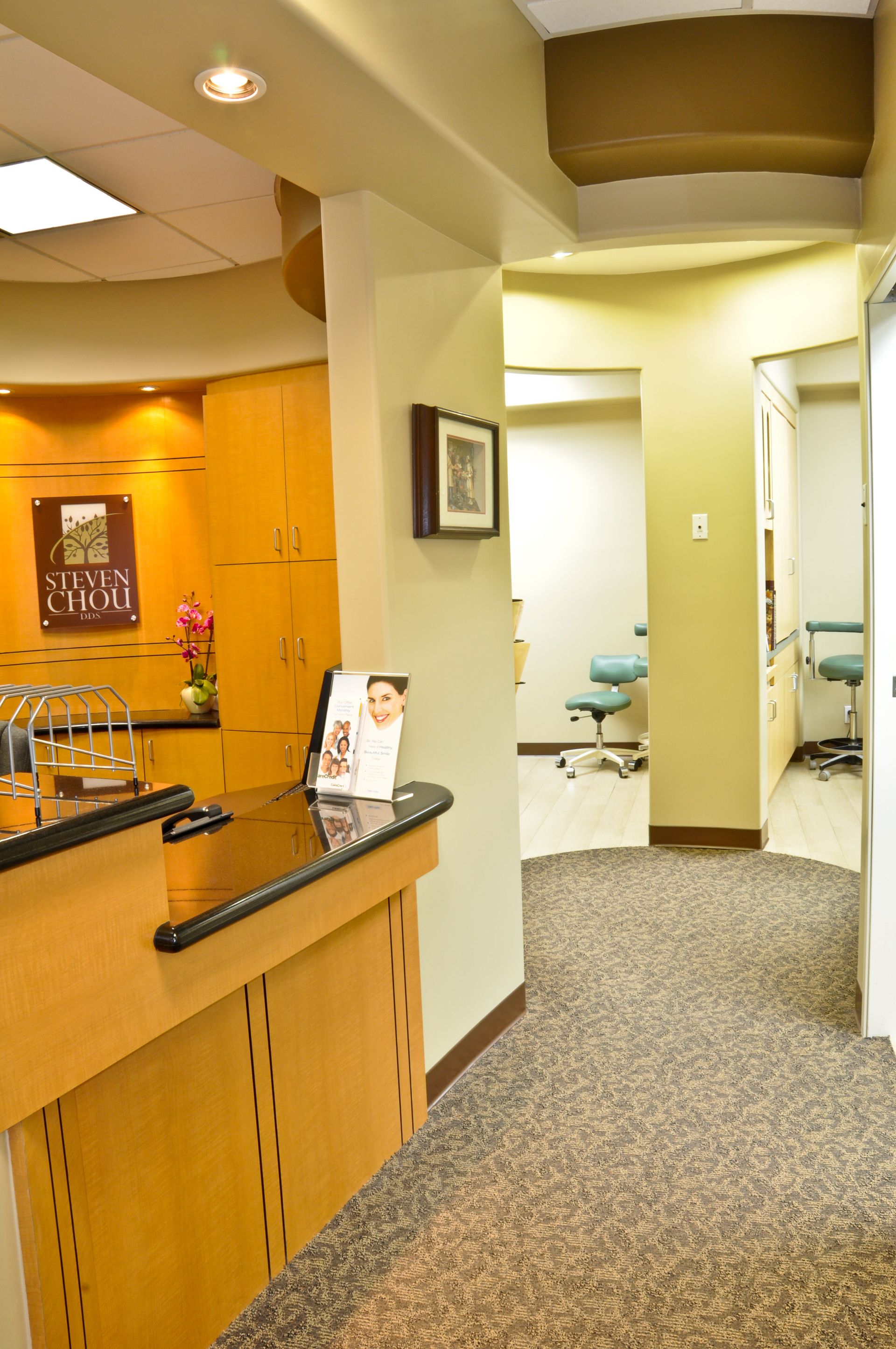 Reception area in a dental clinic with a hallway leading to treatment rooms; beige and brown tones.