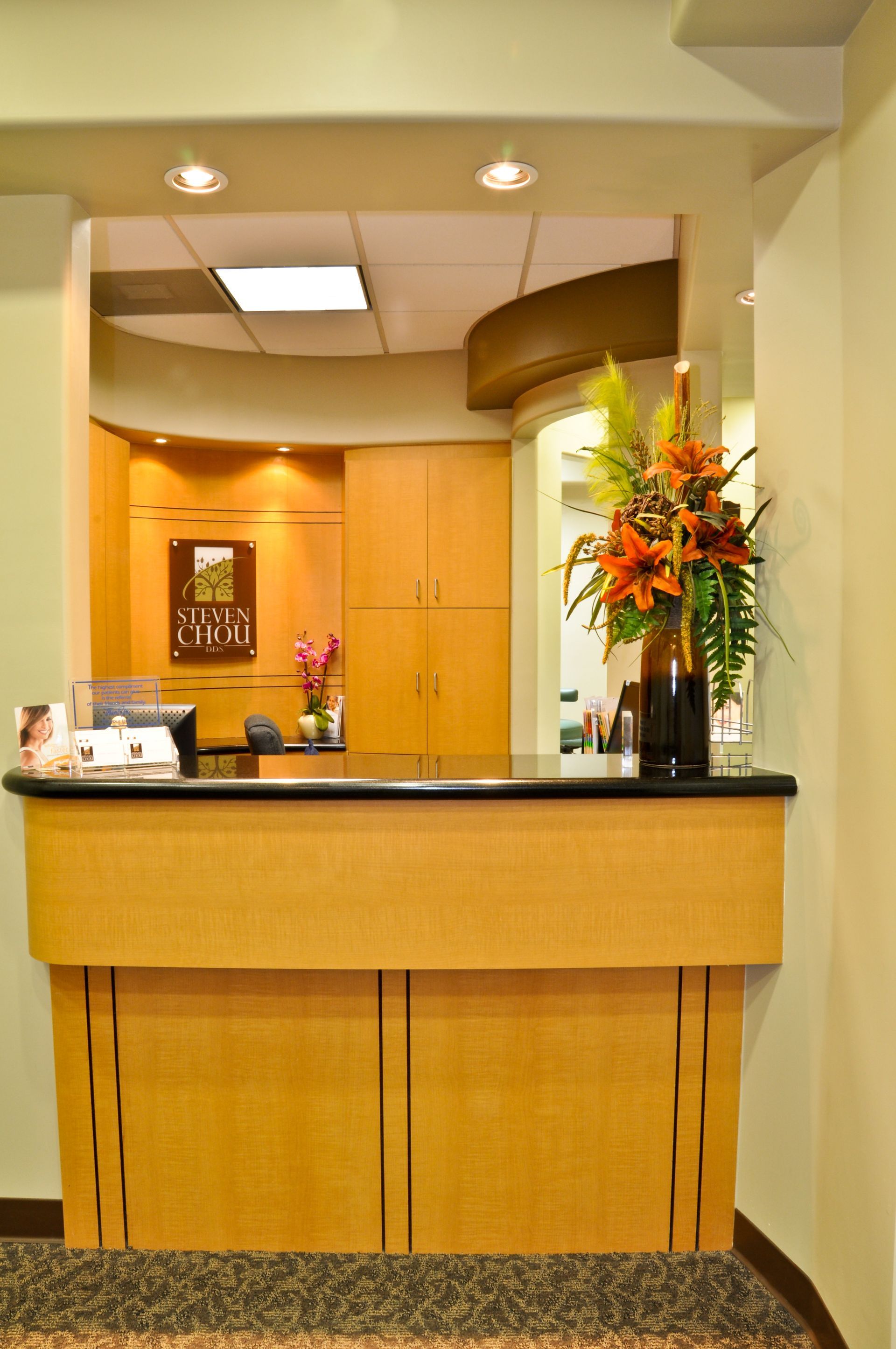 Reception desk in a medical office, beige wood with a dark counter, flowers, and a window to the back office.