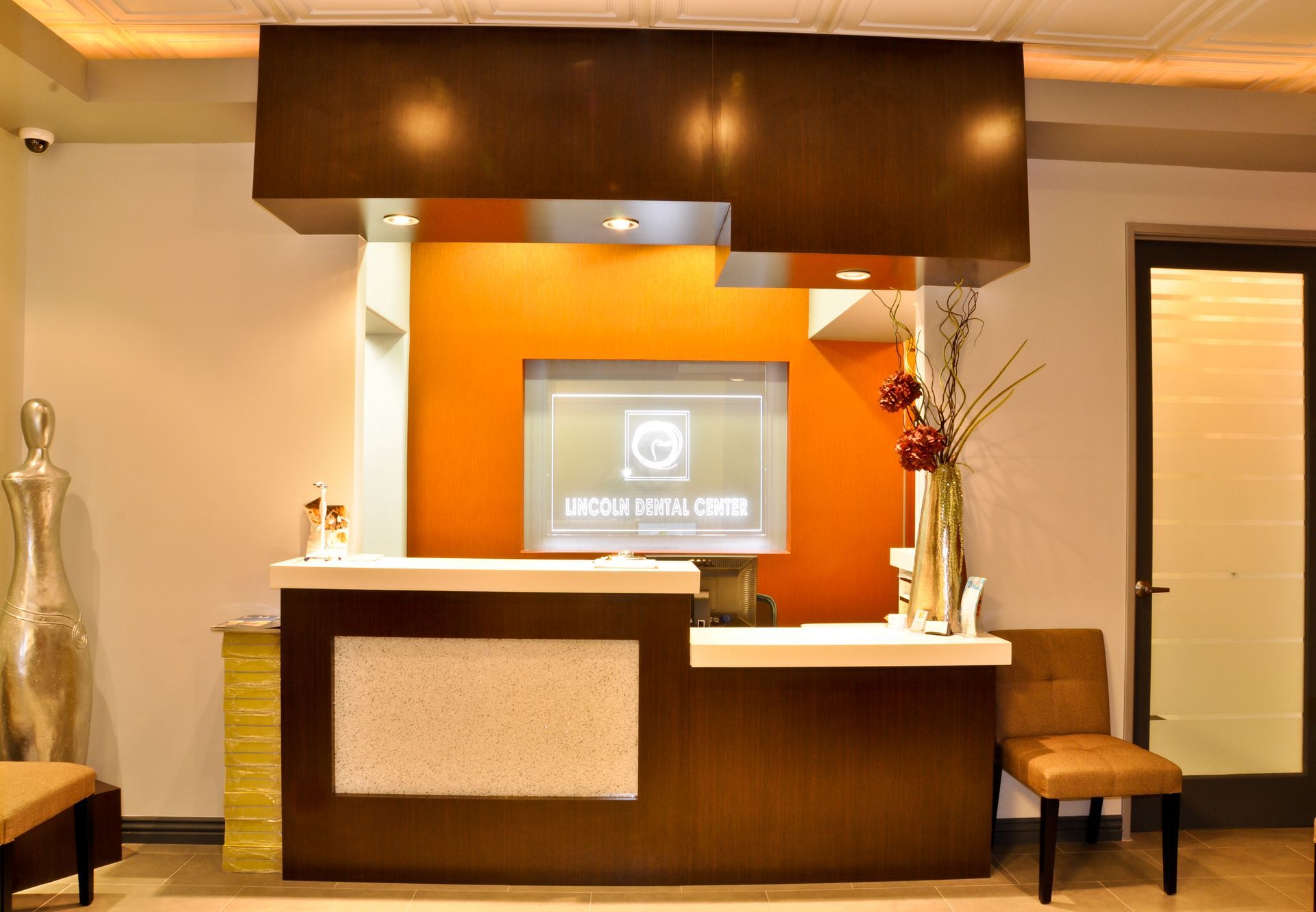 Reception desk in a clinic; brown and white with orange accent wall, a digital display, and a chair.