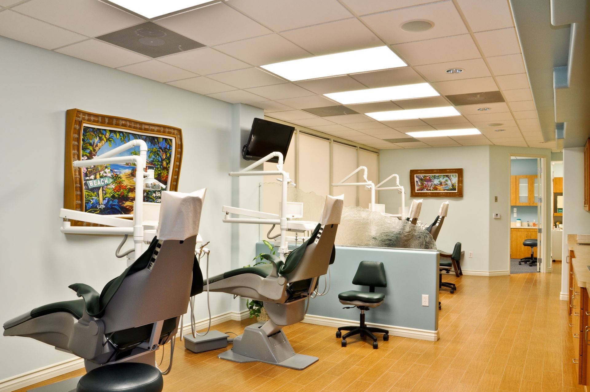 Dental office with chairs, equipment, and artwork. Pale blue walls, wood floors, bright lights.
