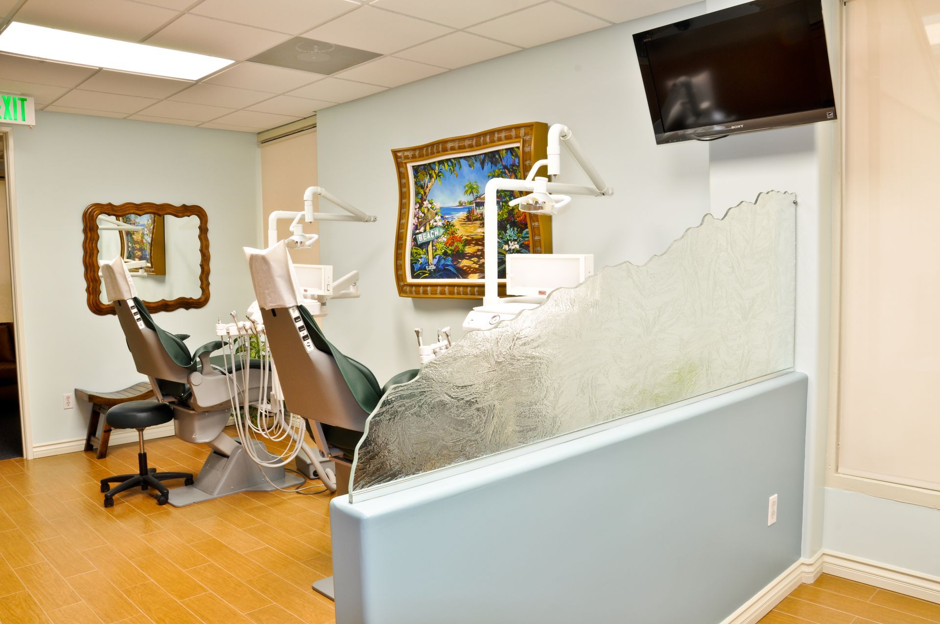 Dental office with two chairs, equipment, art, and a partial frosted glass barrier.