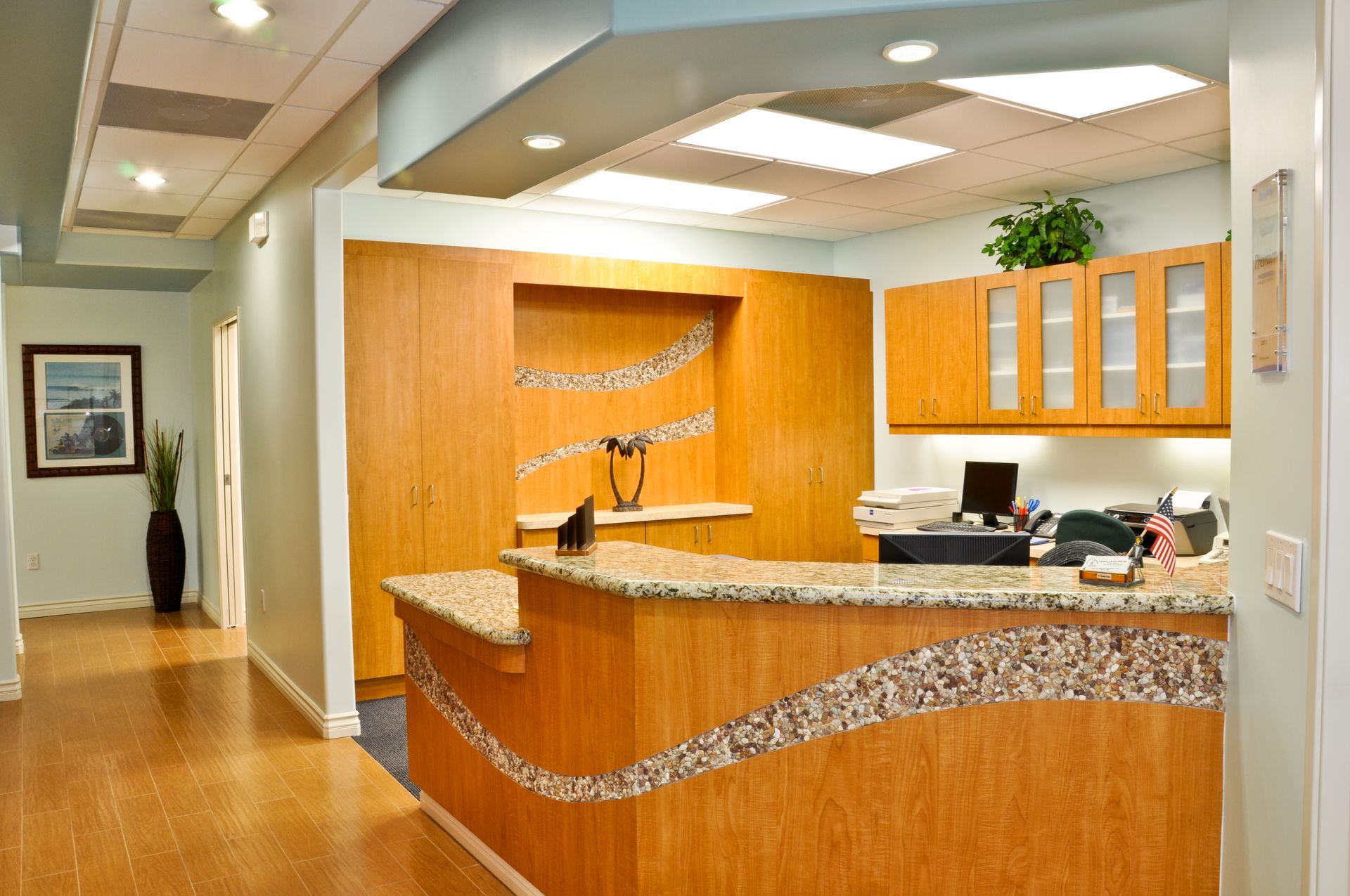 Reception desk in a light wood and stone pattern with a computer, printer, and framed art; neutral colors.