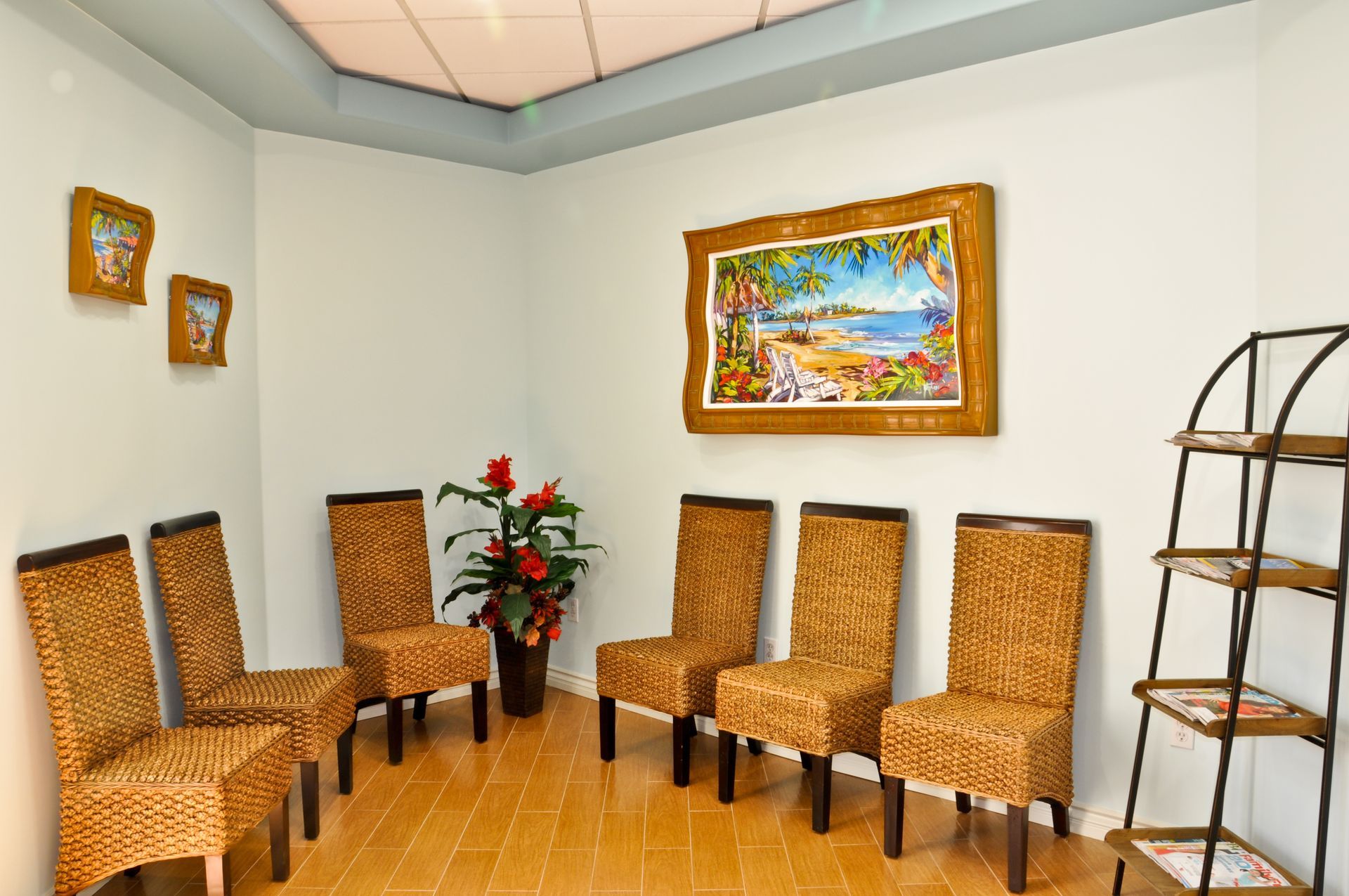 Waiting room with six woven chairs, a coastal painting, and a bookshelf.