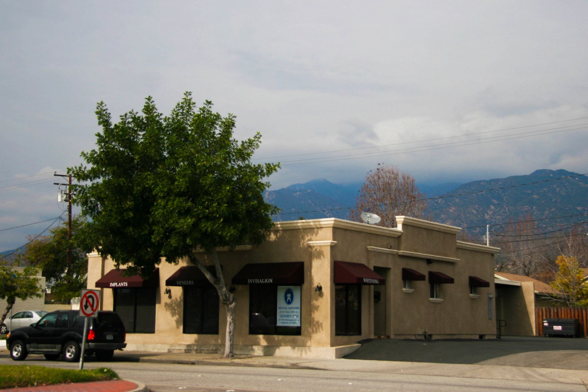 Tan building with awnings, tree, parked car, and mountain backdrop under cloudy sky.
