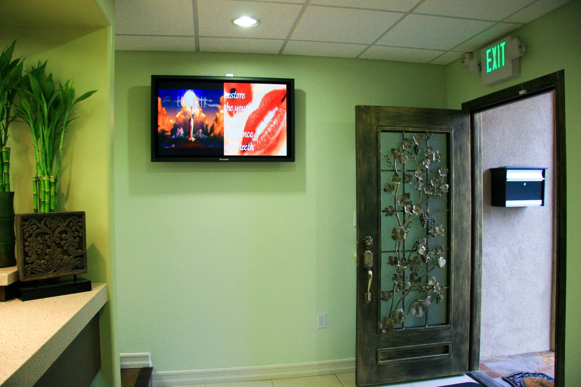 A green-walled room with a TV, an open ornate door, and a desk with plants.