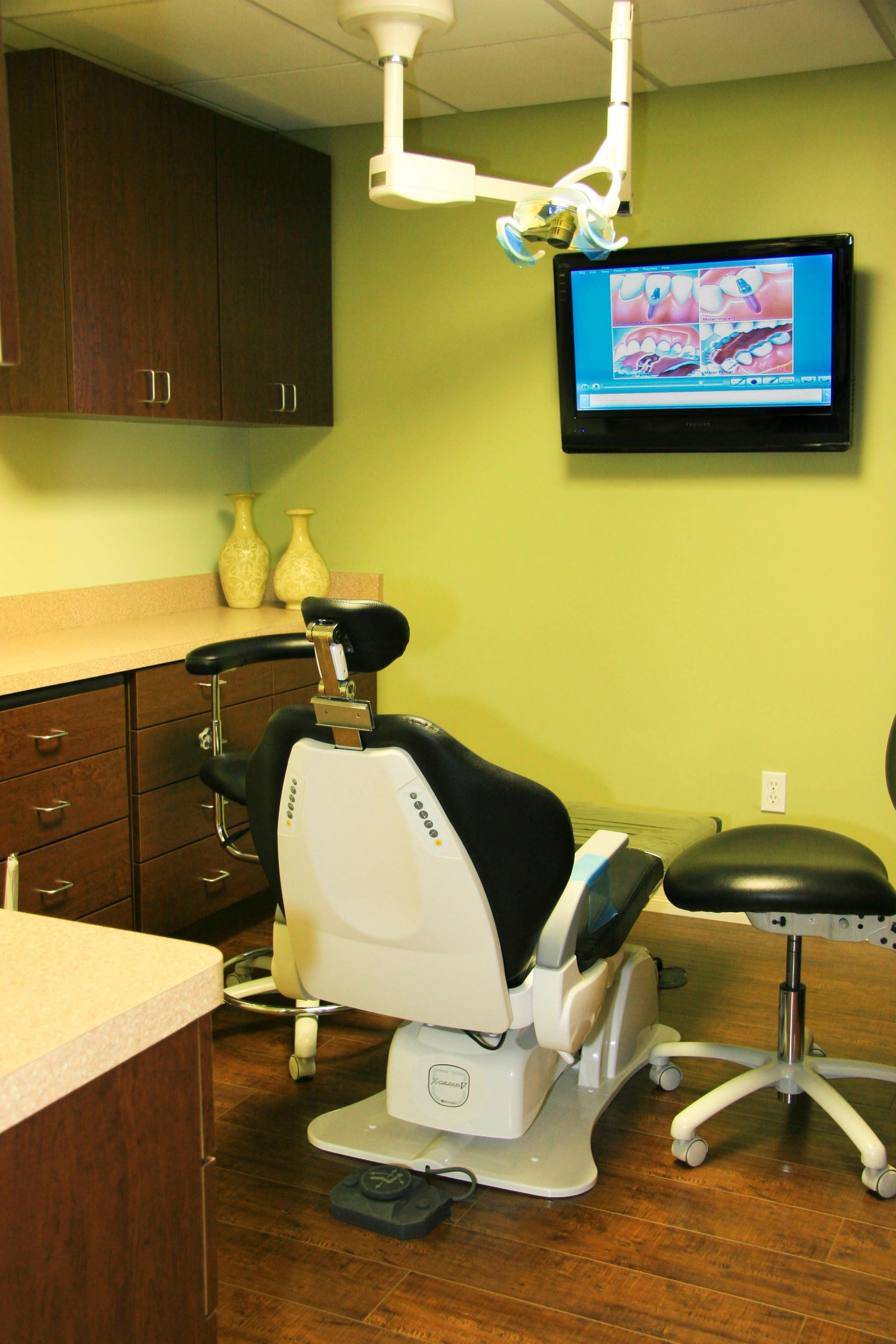 Dental office examination room: dental chair, cabinets, monitor displaying tooth cross-section, green walls.
