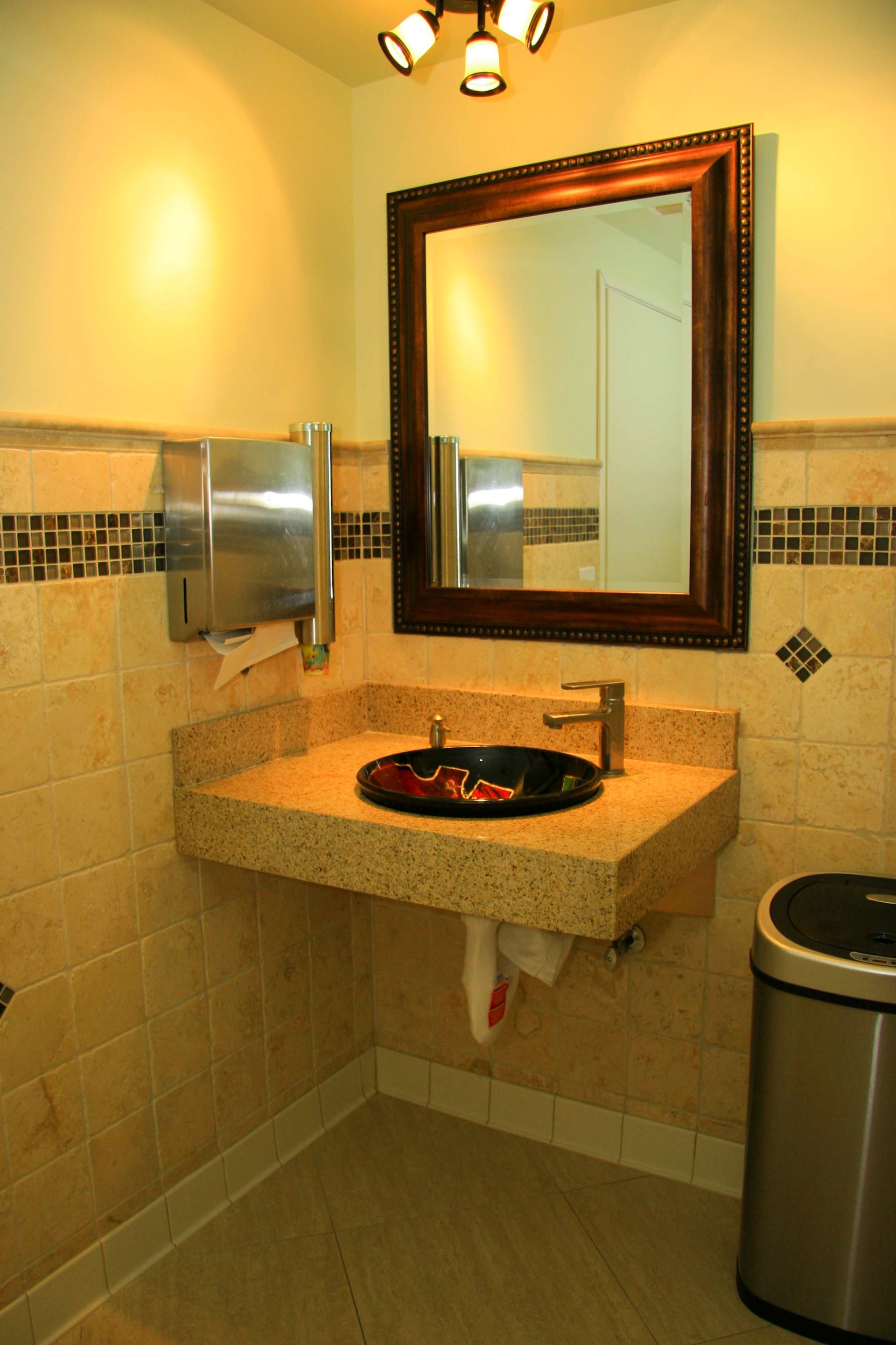 ADA-compliant restroom with a brown-framed mirror, black sink, granite countertop, and a stainless steel paper towel dispenser.