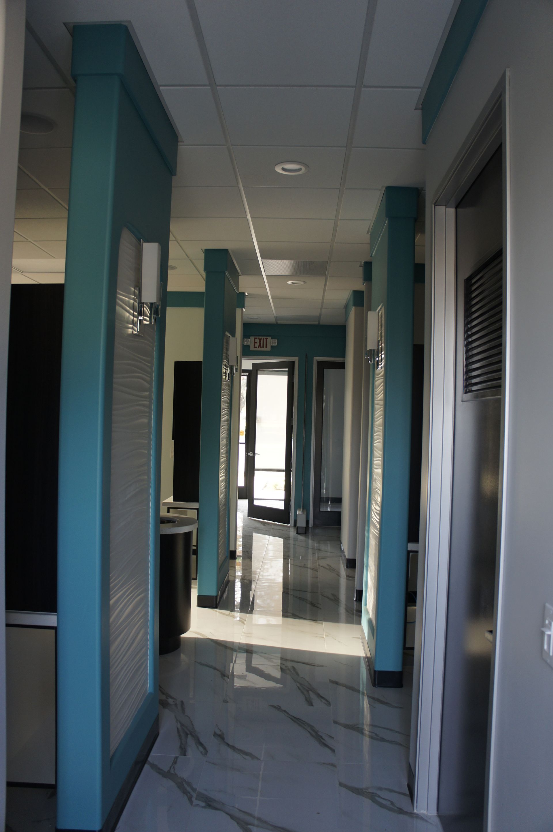 Hallway with teal pillars, marble floor, and doors leading to rooms; sunlight at the end.