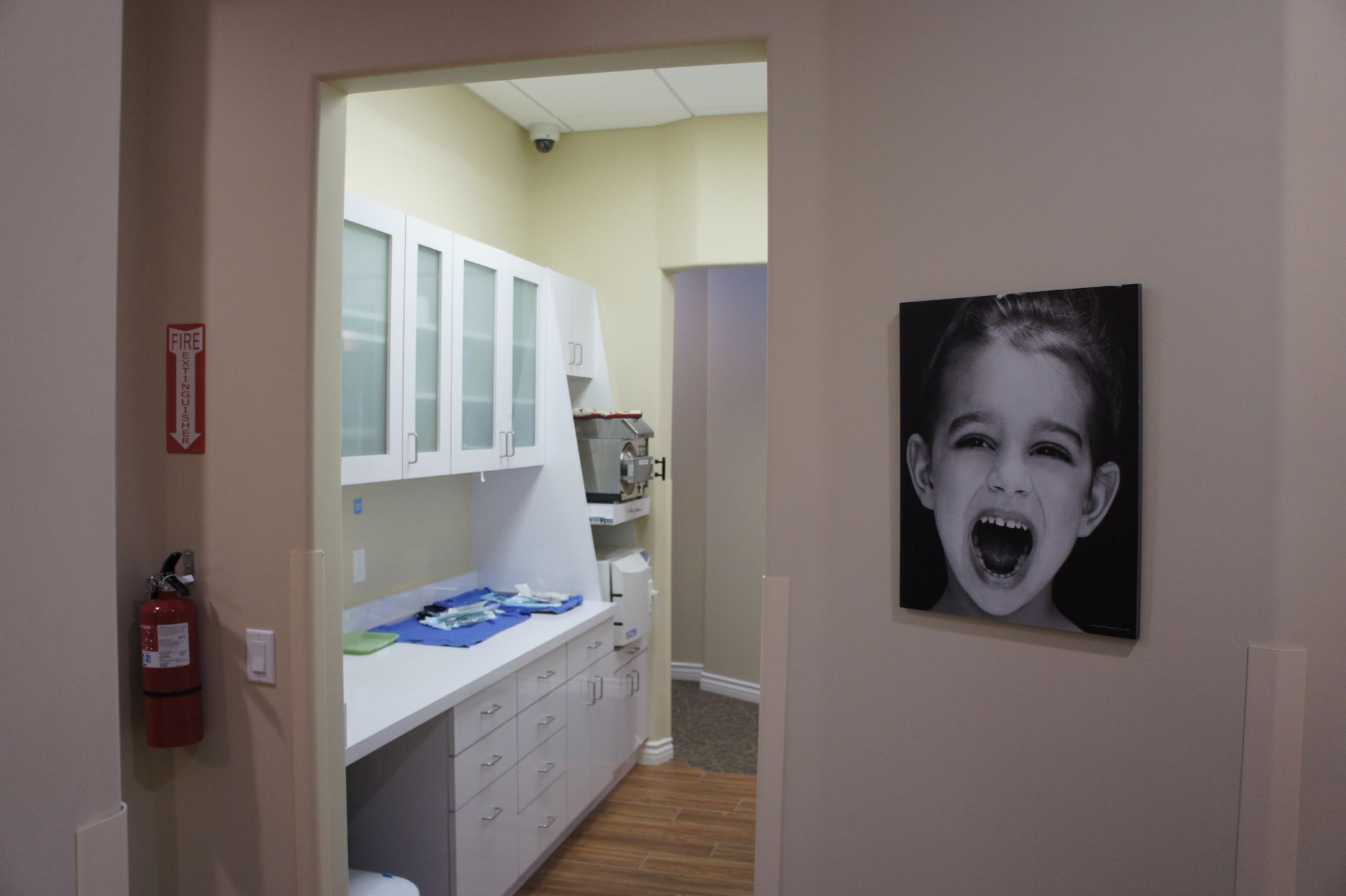 Hallway with a dental office exam room visible, featuring cabinets, counter, equipment, and child’s portrait.