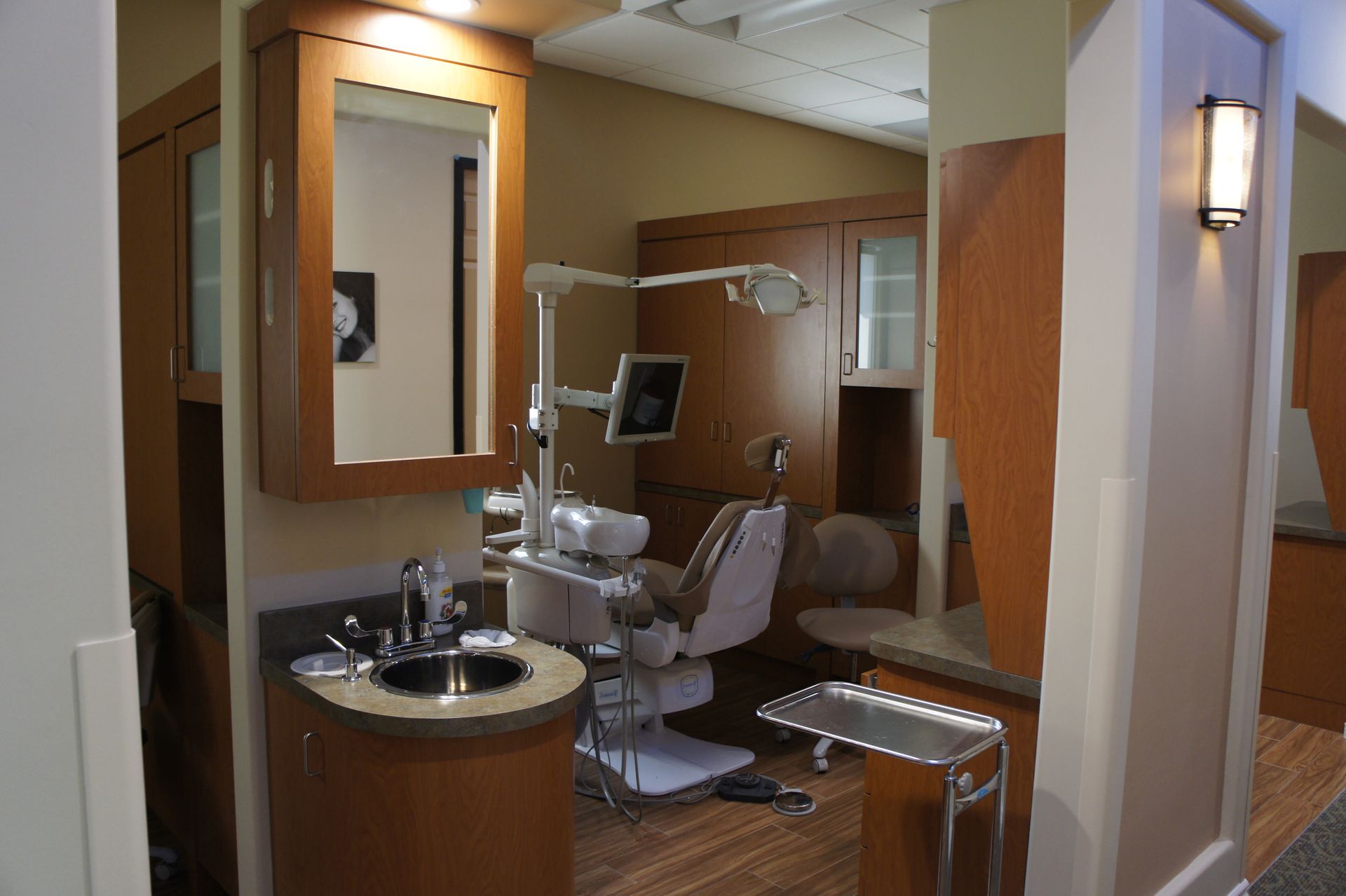 Dental operatory room: chair, cabinets, mirror, sink, dental light; light wood tones.